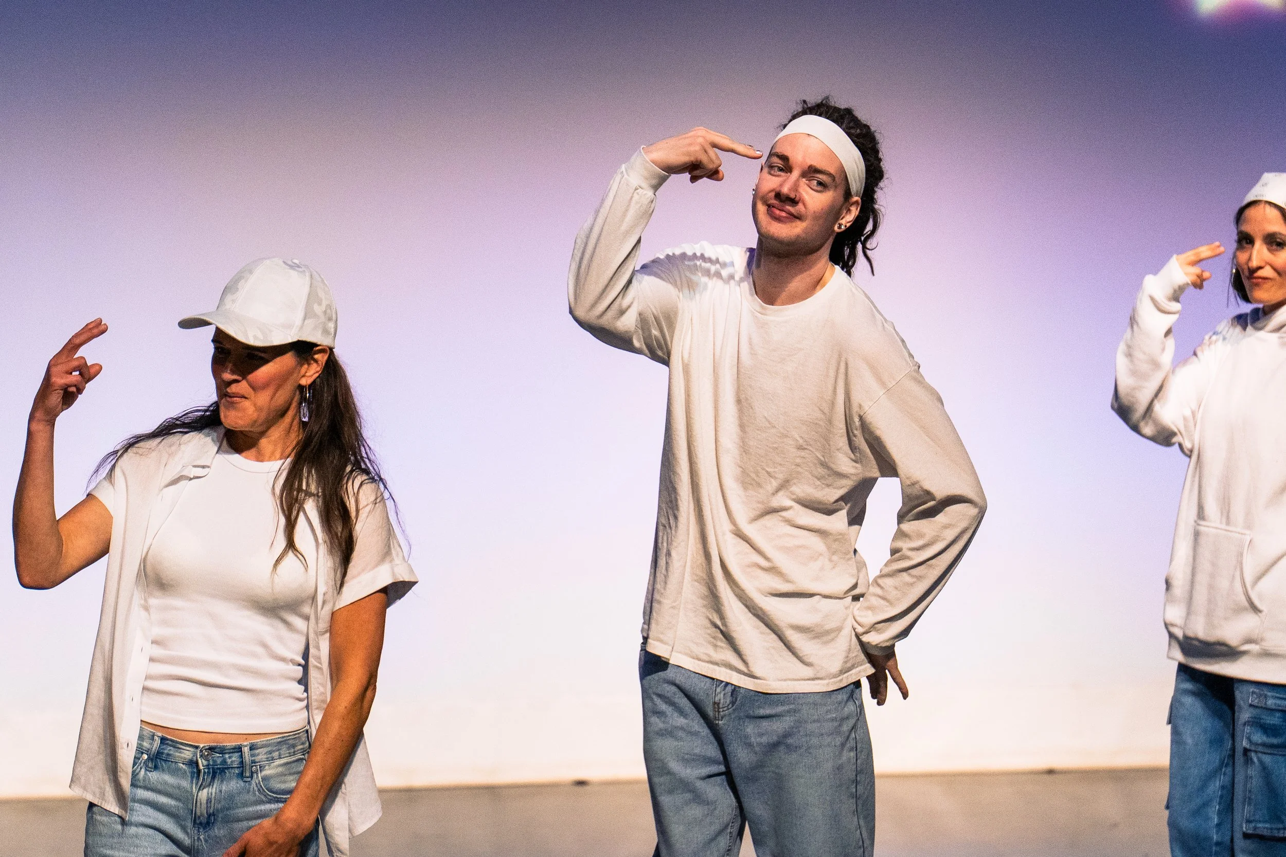 Three performers on stage in casual white clothing, making hand gestures near their heads, with a gradient purple-to-white background.