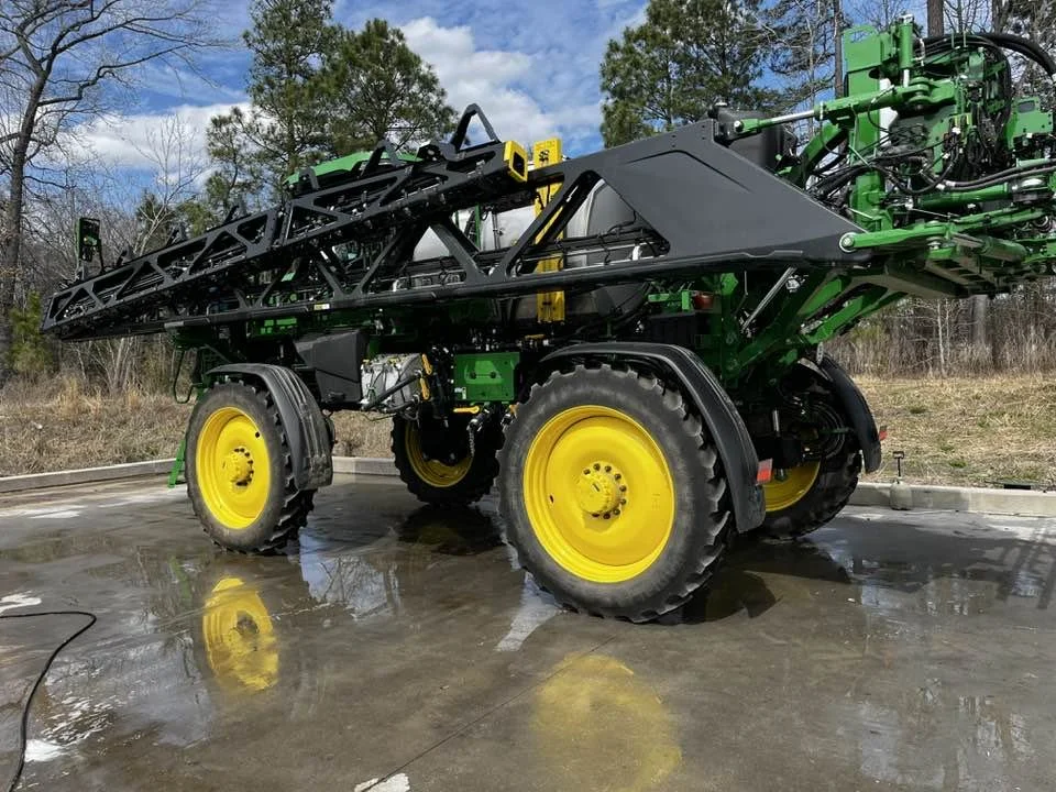 Agricultural sprayer with large yellow wheels on a concrete surface, surrounded by trees.