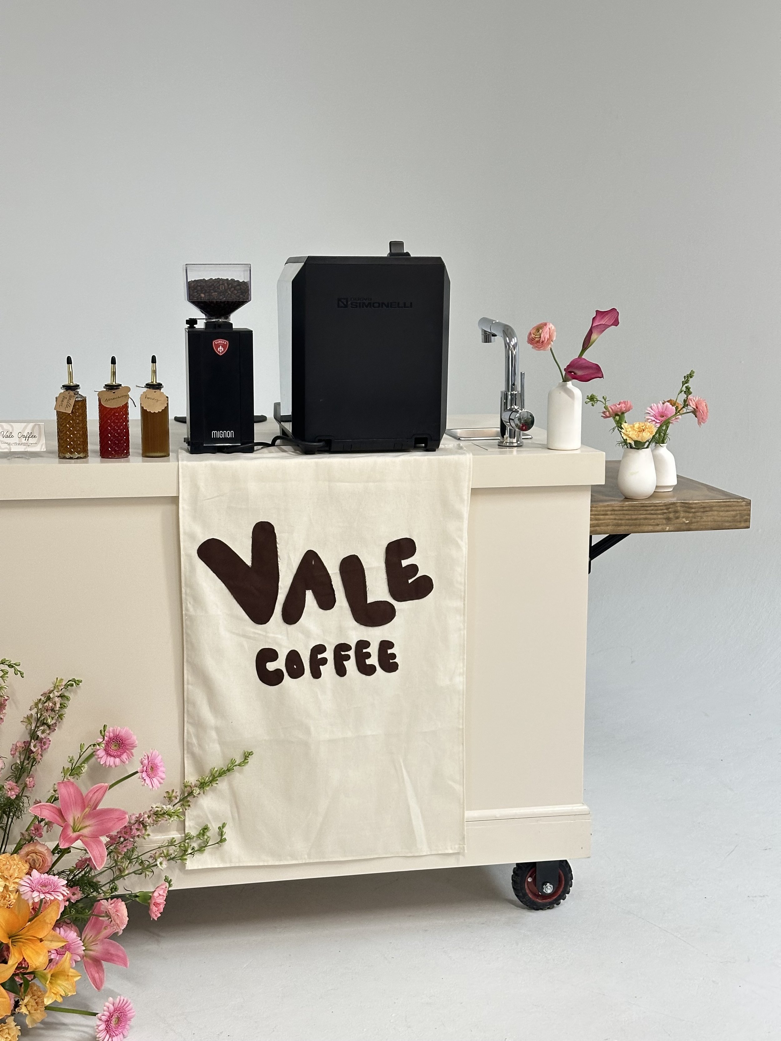 A coffee station with a white cloth hanging from a cart labeled "Vale Coffee." The cart has coffee brewing equipment, including a bean grinder and a machine. There are jars with flavored syrups to the left and floral arrangements in white vases on th