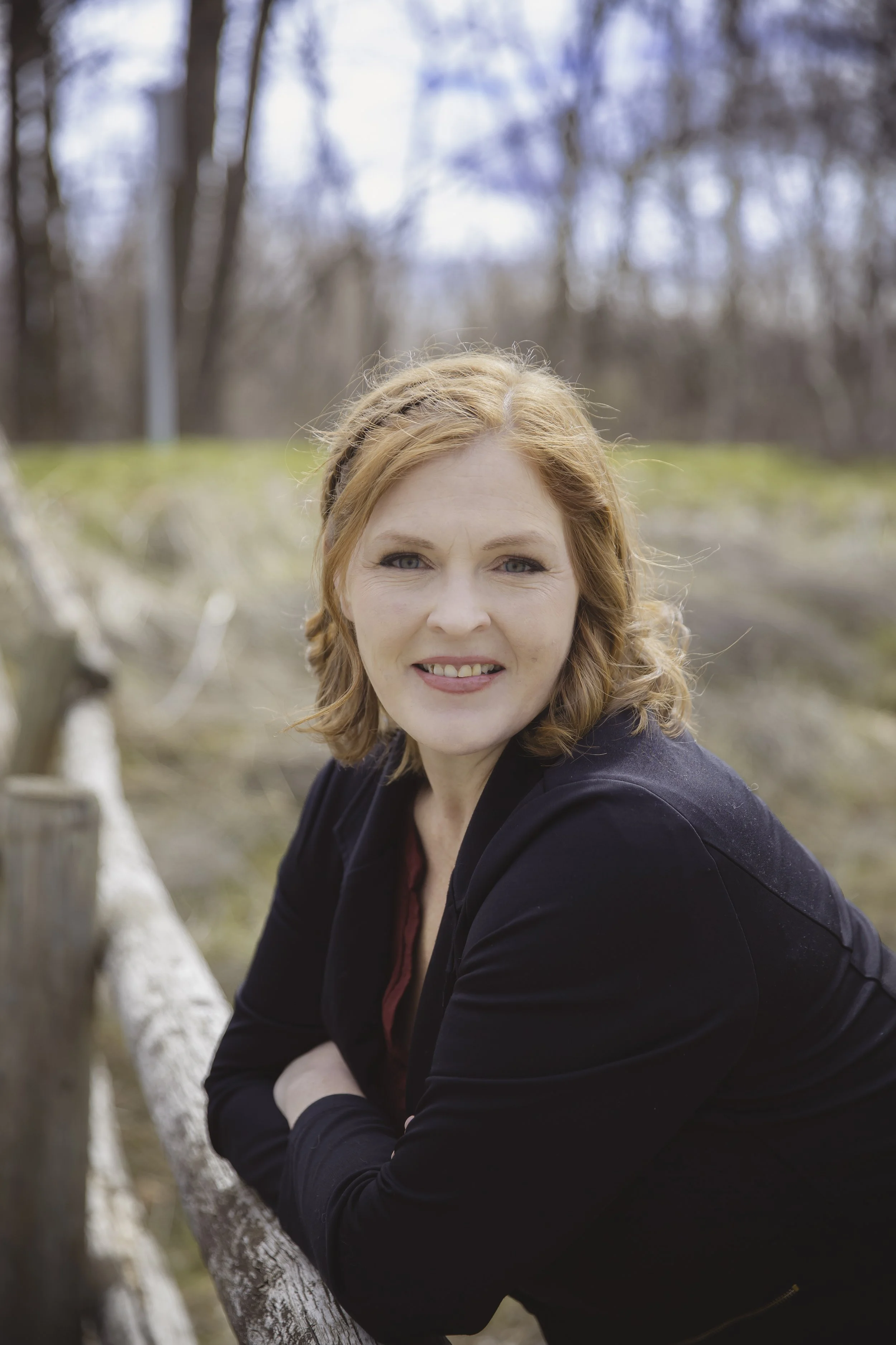 A woman with red hair leaning on a wooden fence outdoors, smiling, surrounded by trees.