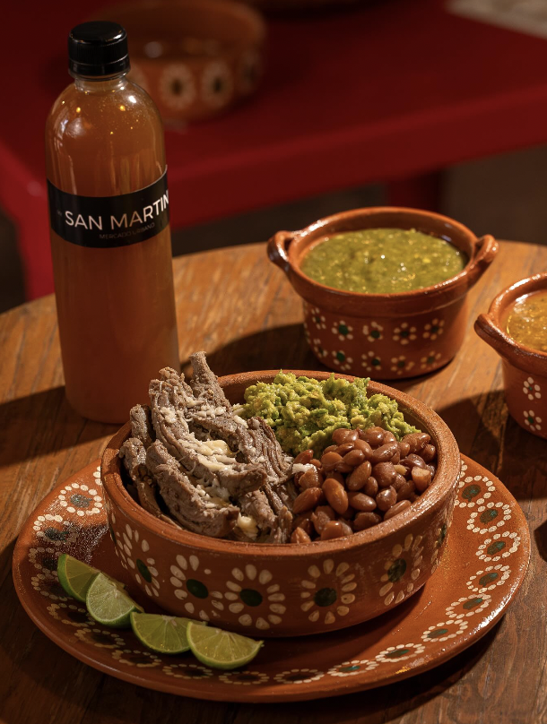 Comida mexicana tradicional con carne deshebrada, frijoles y guacamole en un tazón de barro decorado, acompañada de rodajas de lima, salsa en pequeños tazones y una bebida embotellada sobre una mesa de madera.