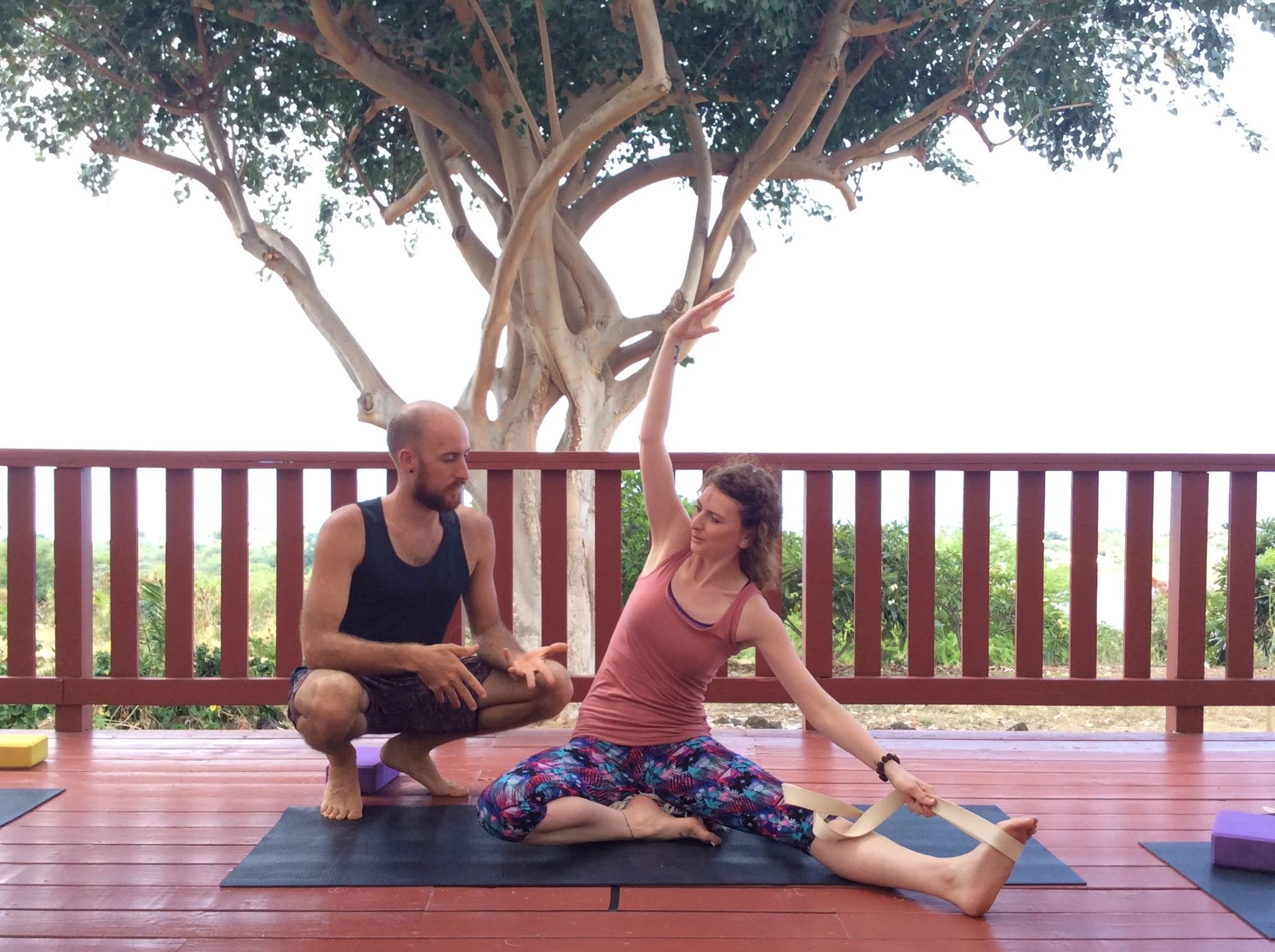A teacher guiding a student through a seated side stretch on a wooden deck.