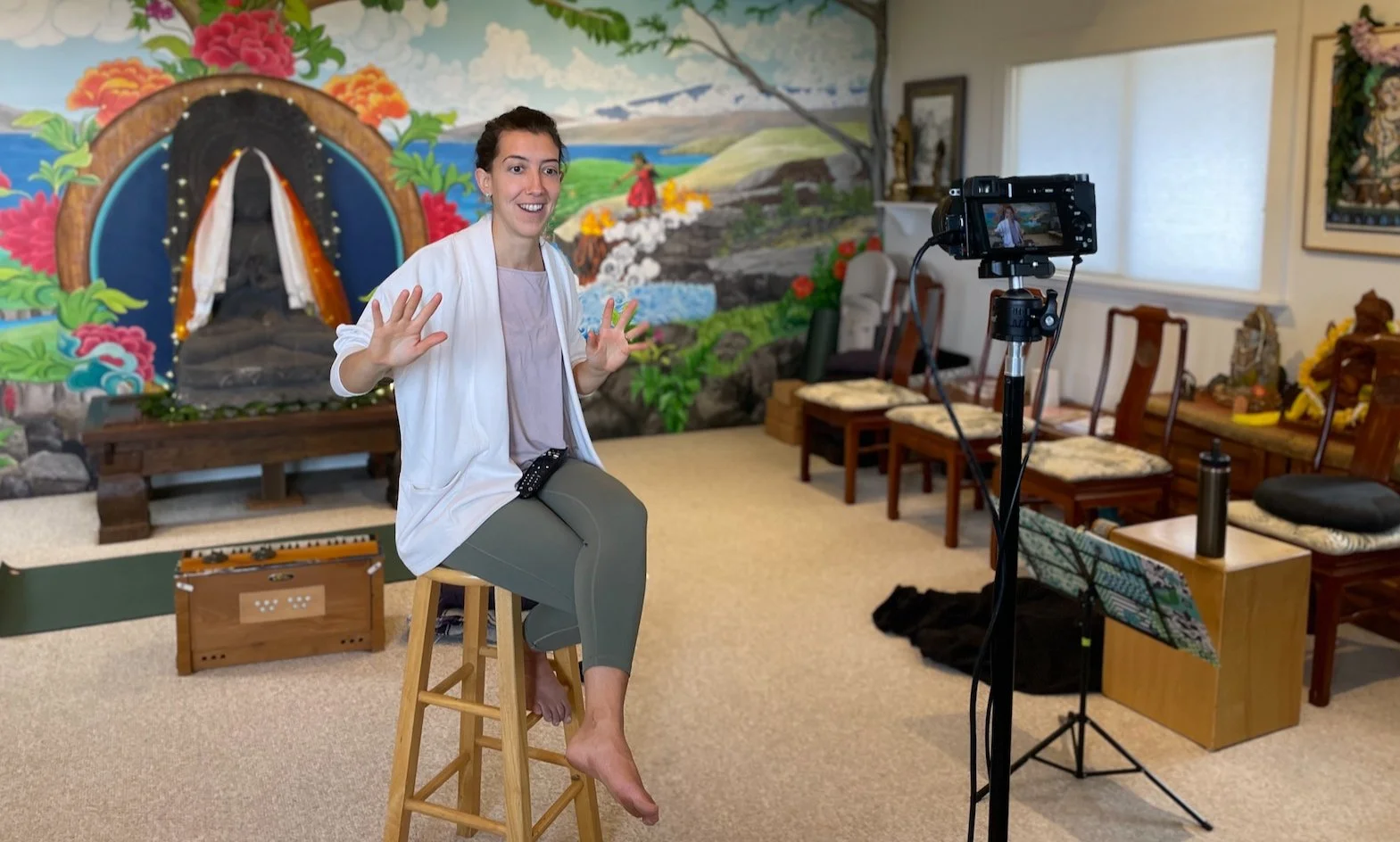 A woman sitting on a stool in front of a camera recording a video in a room with a colorful mural of a mountain landscape and a shrine background.