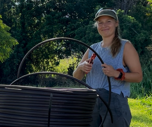 Young girl outdoors holding watering tool, standing next to a black garden hose reel.
