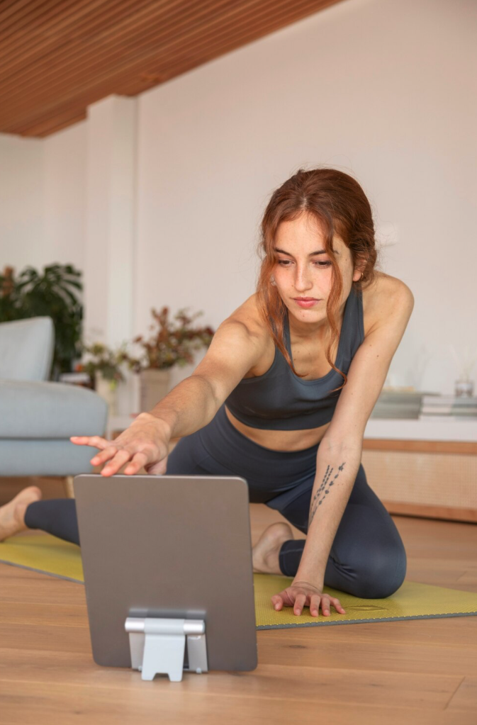 A woman following an online yoga class on a laptop beside her mat.