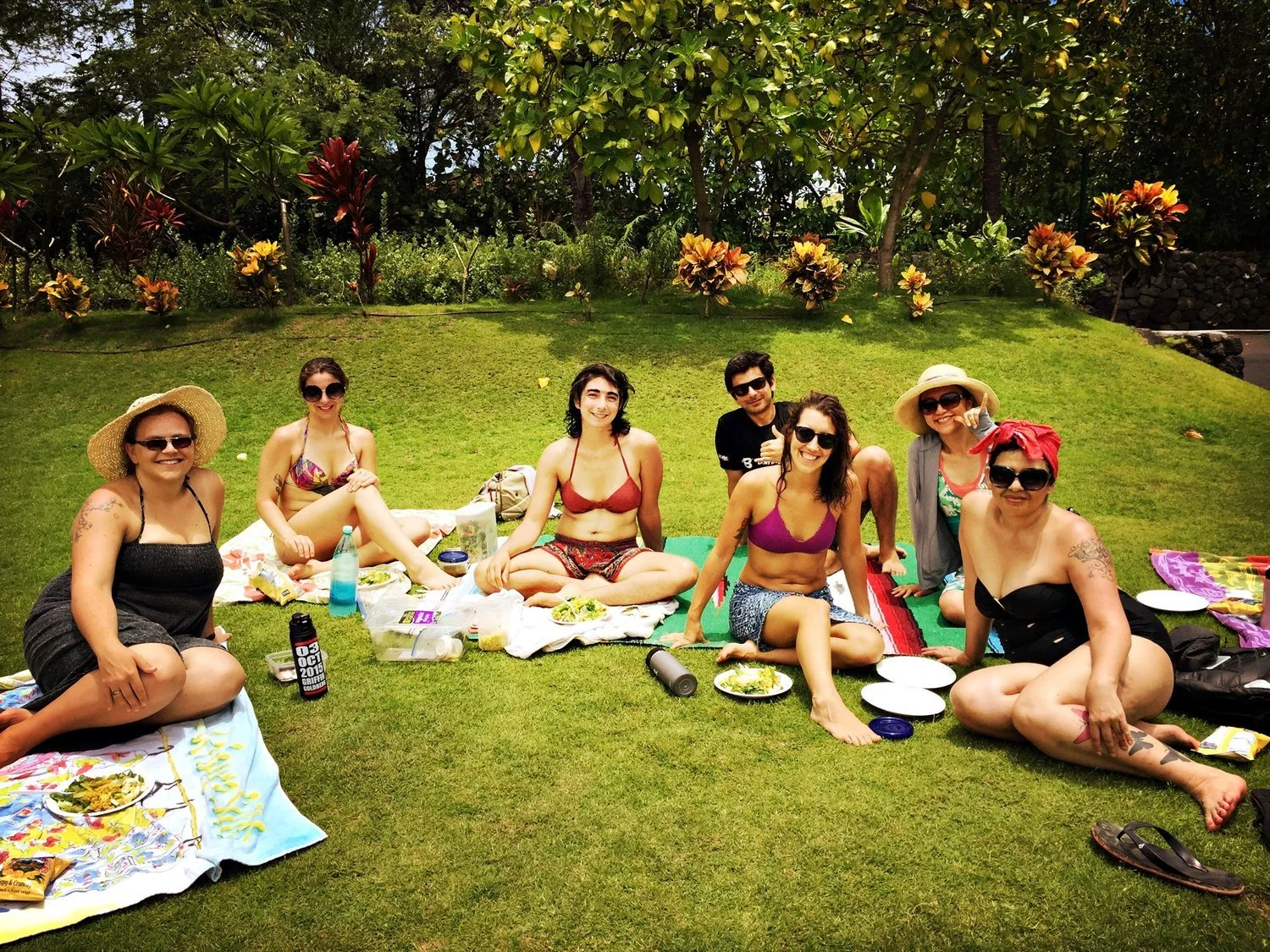 Group of friends enjoying a picnic on a grassy lawn with trees and colorful plants in the background.