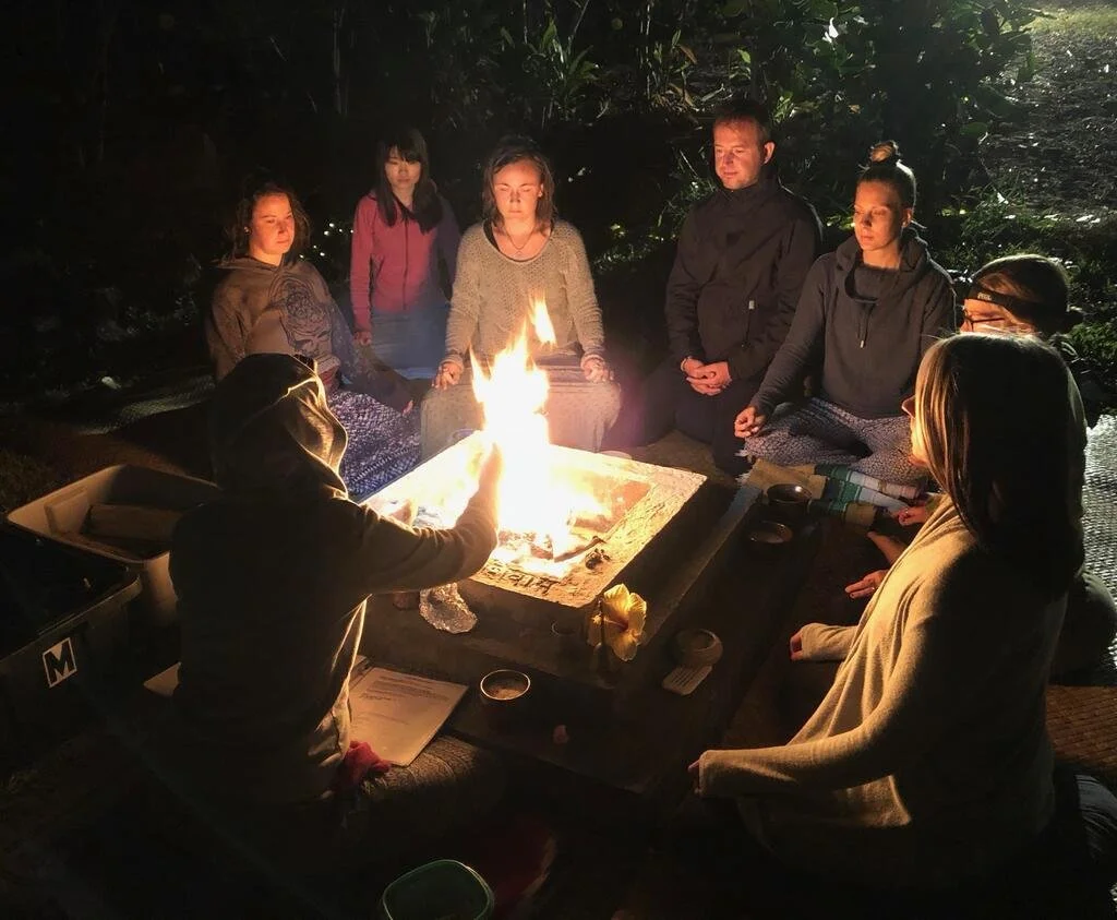 A group of yogis sit cross legged around a fire in the early morning.