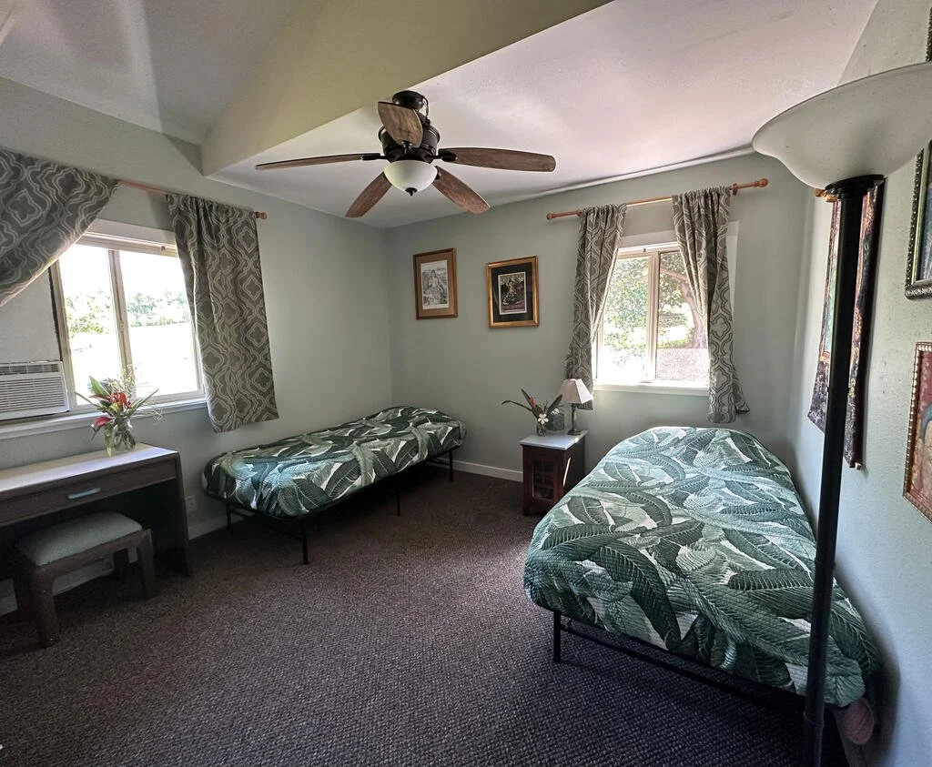 A pristine shared room with two beds, tropical patterned comforters, a fan in the ceiling, and art on the walls.