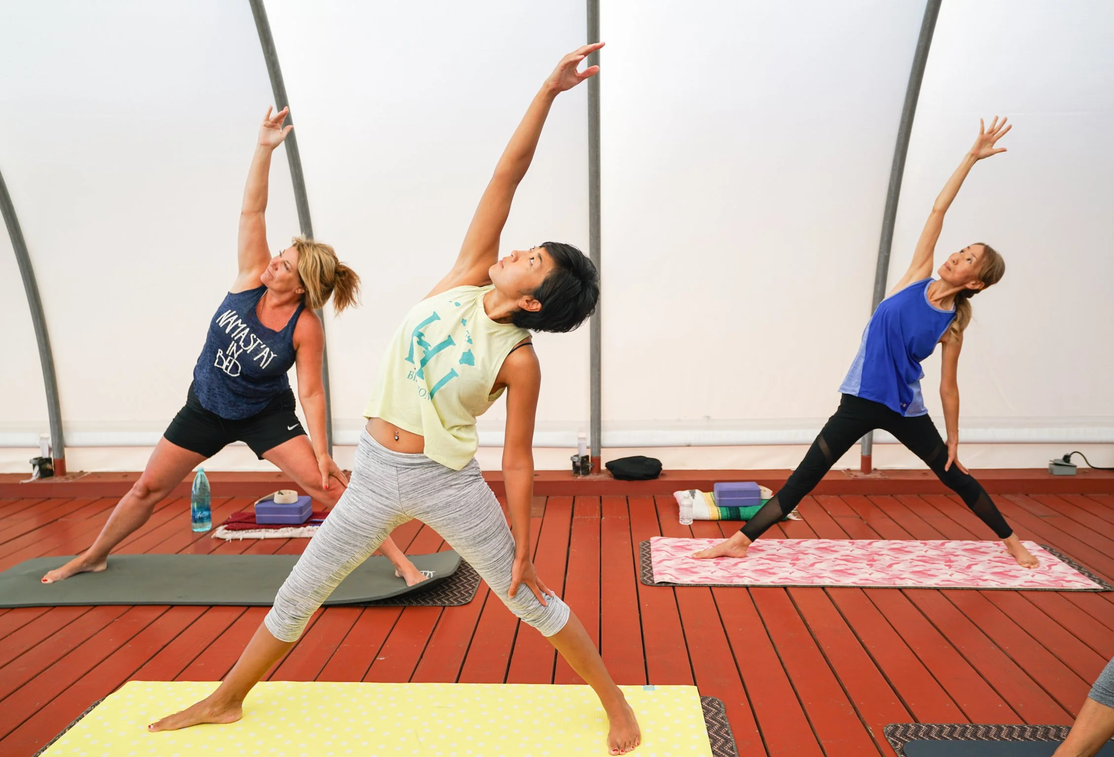 Three women practicing yoga indoors on mats, doing wide-legged forward bends with arms reaching upward.