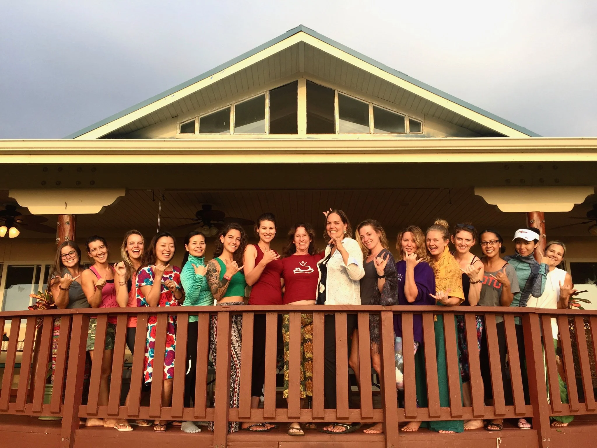 Group of women and girls standing on a wooden balcony in front of a house, smiling and making hand gestures at sunset.