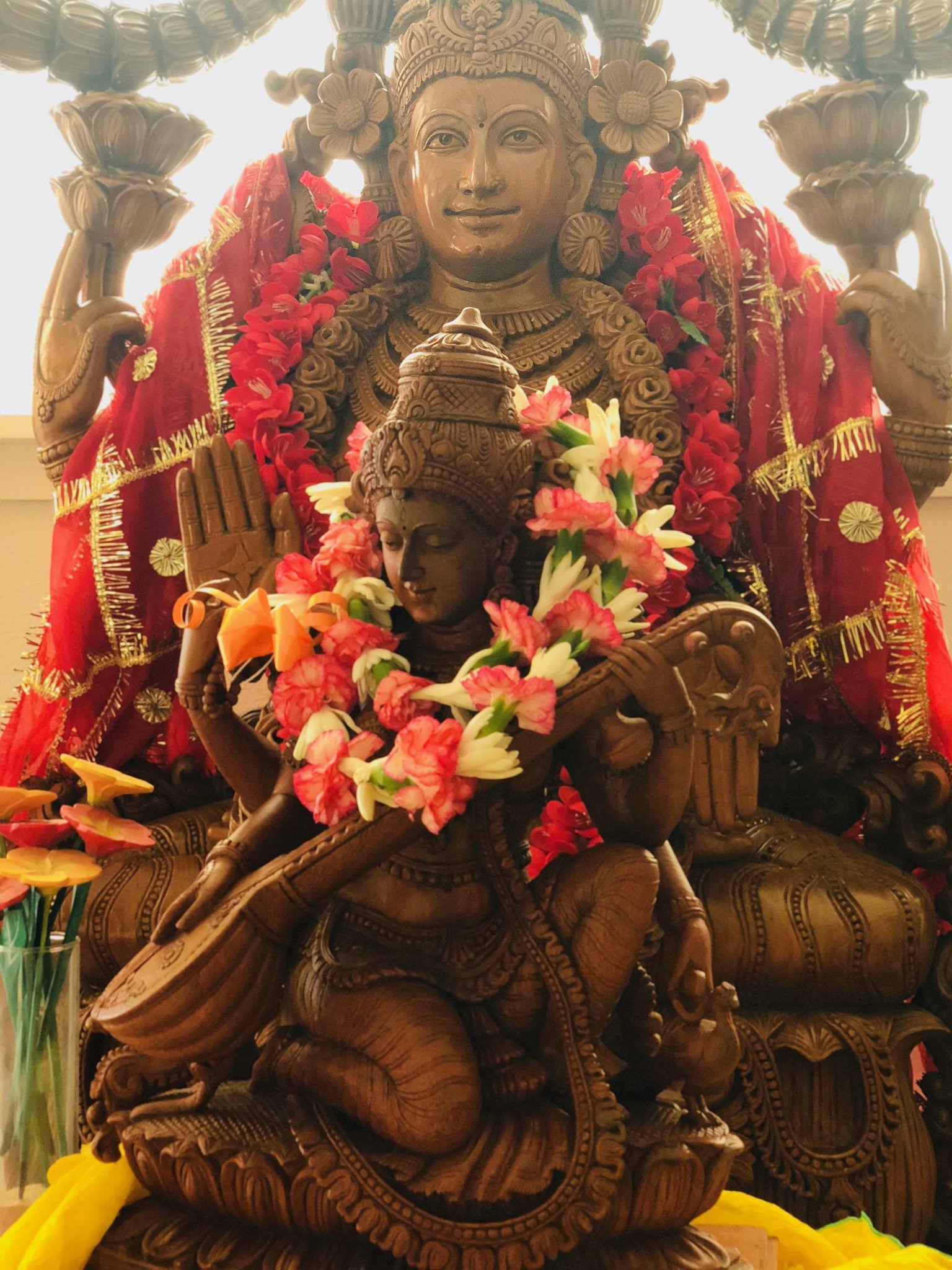 Wooden statue of a Hindu deity adorned with red and gold cloth, floral garlands, and jewelry, with intricate carvings and a seated posture.
