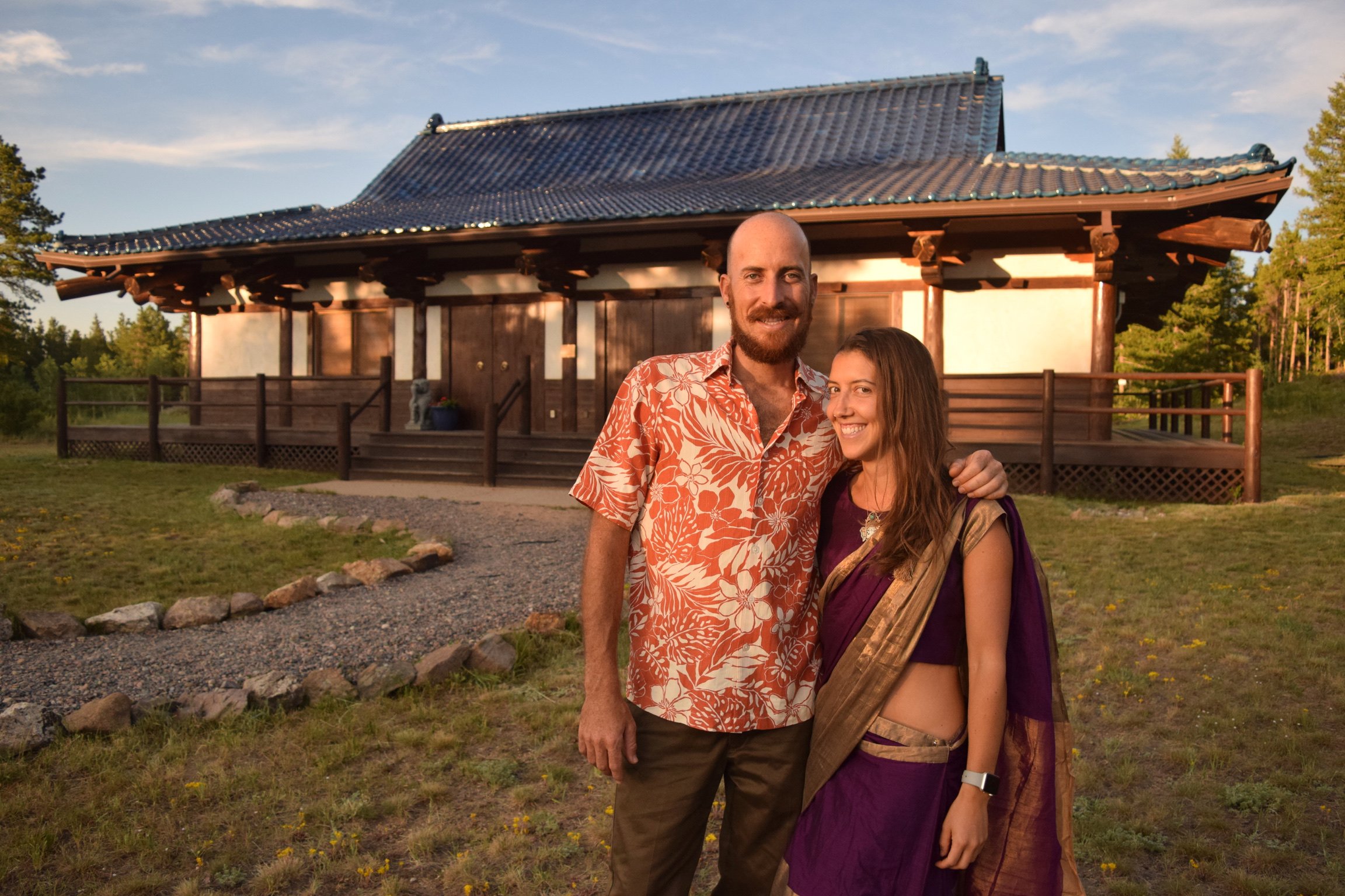 Two yoga instructors smiling and standing close together outside a traditional Japanese-style temple, with greenery and trees in the background during sunset.