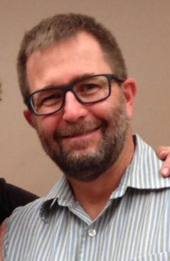 A smiling man with glasses, short brown hair, a beard, wearing a striped shirt.