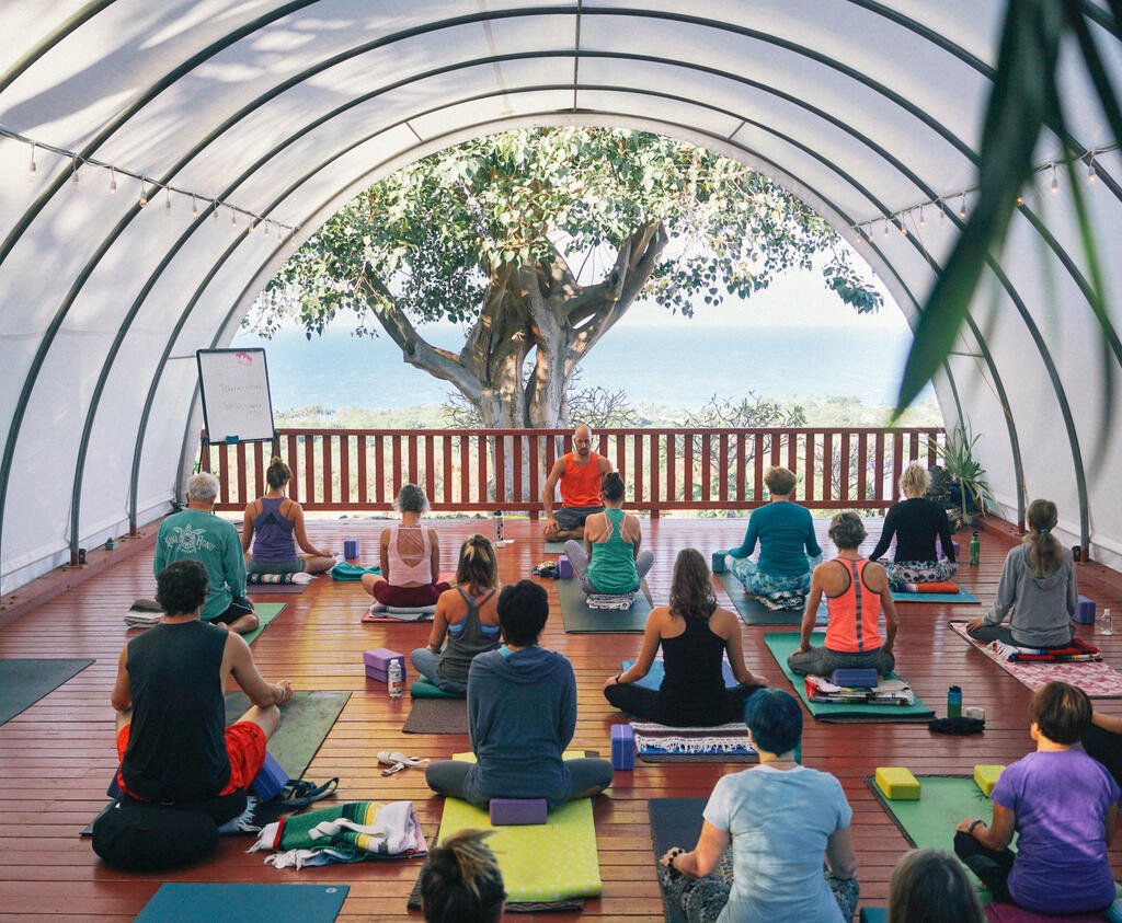 A group of yogis sit cross-legged in front of an instructor on a covered deck overlooking the ocean.