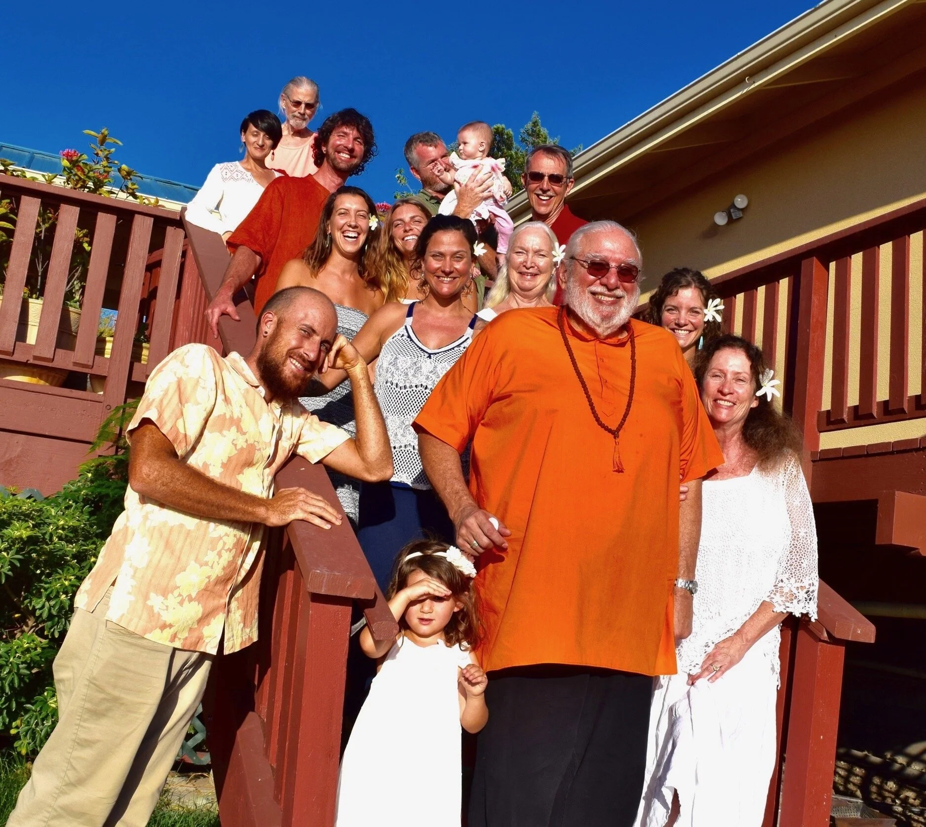 A group of people, including children and adults, smiling and gathered on a wooden staircase in front of a yellow house with a blue sky background, possibly at a celebration or family gathering.