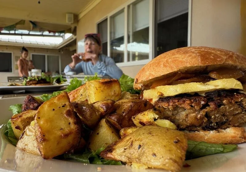 Close-up of a cheeseburger with potato wedges on a plate, with people dining in the background.
