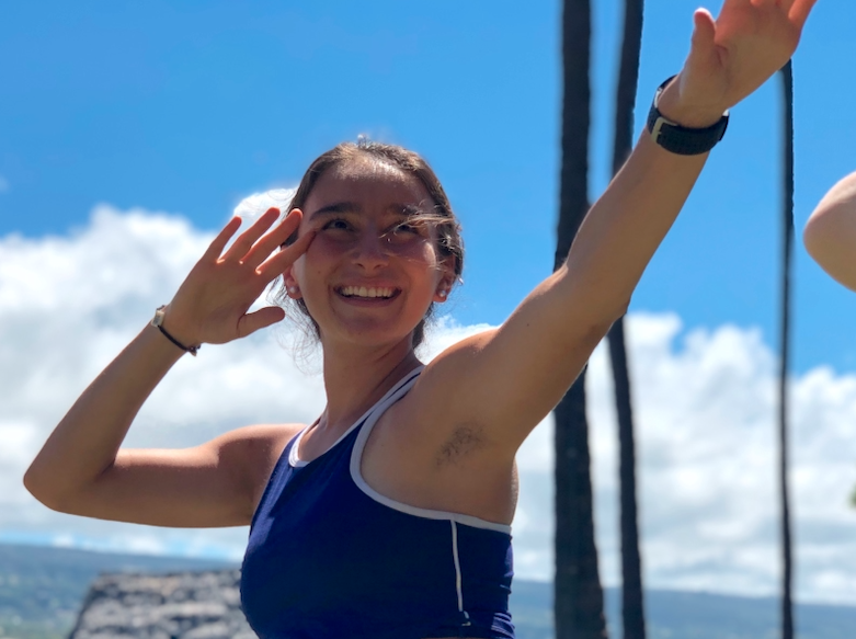 Smiling woman with long hair, wearing a blue athletic top, saluting with her right hand, outdoors with blue sky and trees in the background.