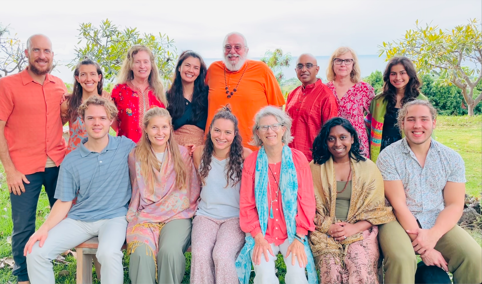 Group of fifteen diverse people outdoors, sitting and standing on grass with green trees and cloudy sky in the background, smiling for a group photo.