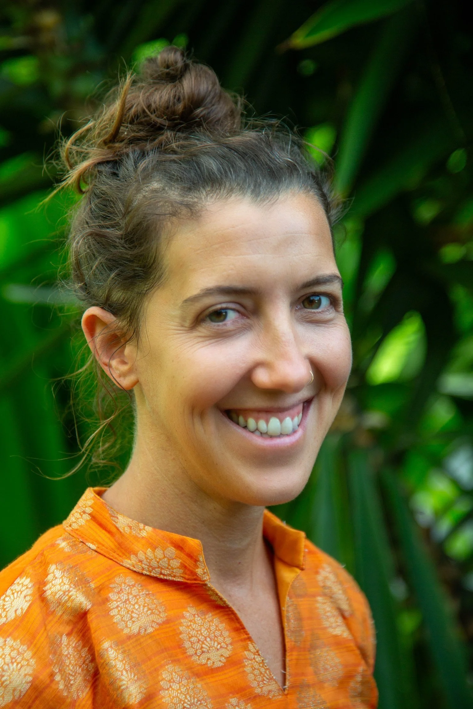 A female yoga instructor with brown hair in a messy bun, wearing an orange shirt with a golden floral pattern, standing outdoors with green foliage in the background.