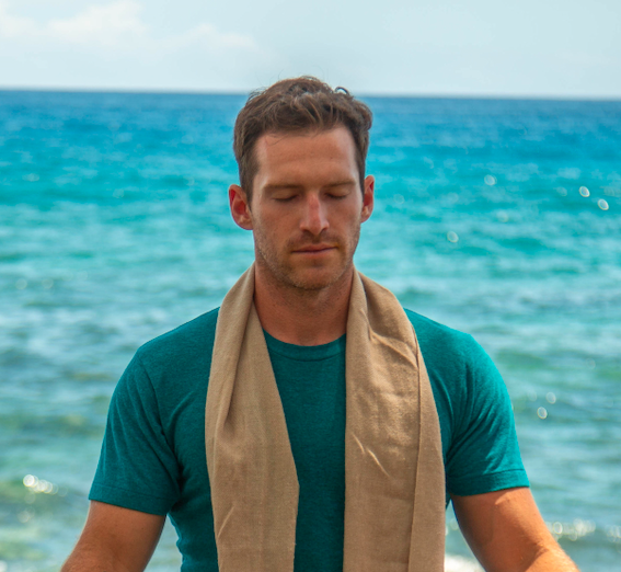 Man in teal t-shirt with beige scarf meditating on the beach with ocean in background.
