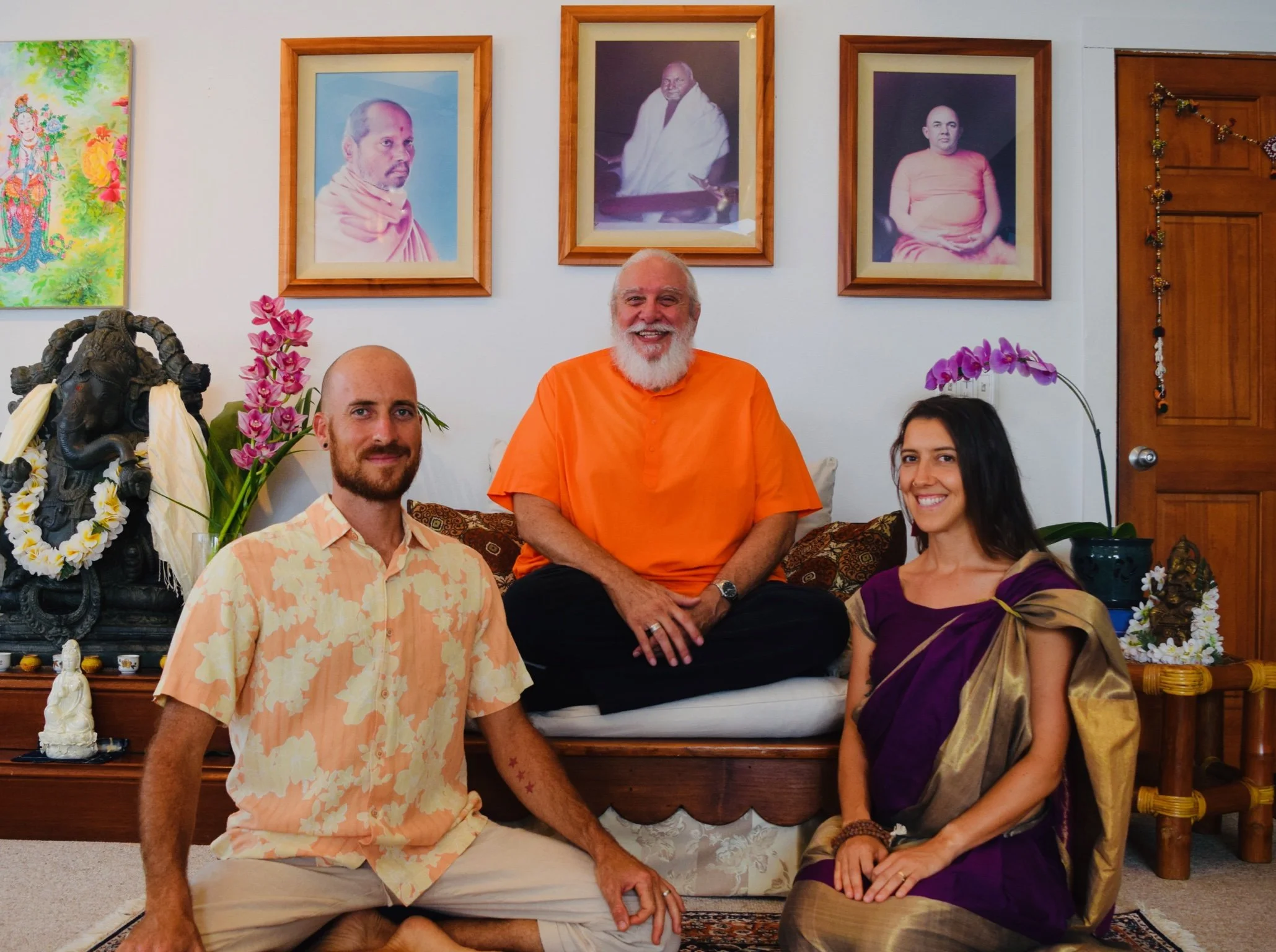 A group of three people sitting indoors in front of framed spiritual portraits, including an elderly man with a white beard in orange robes, a man with a beard in a floral shirt, and a woman in a purple and gold traditional outfit, surrounded by decorative plants and religious statues.
