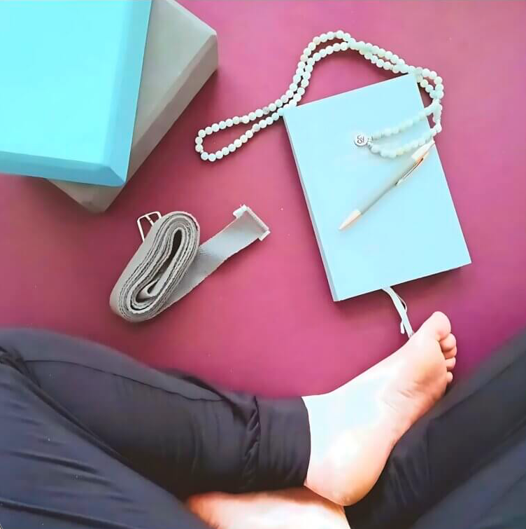 A person's feet and lap are visible, with a pink surface showing. On the surface, there is a blue notebook with a pen, a white beaded necklace, a rolled-up strap, and some boxes in blue and beige. The scene appears to be a workspace or sitting area.
