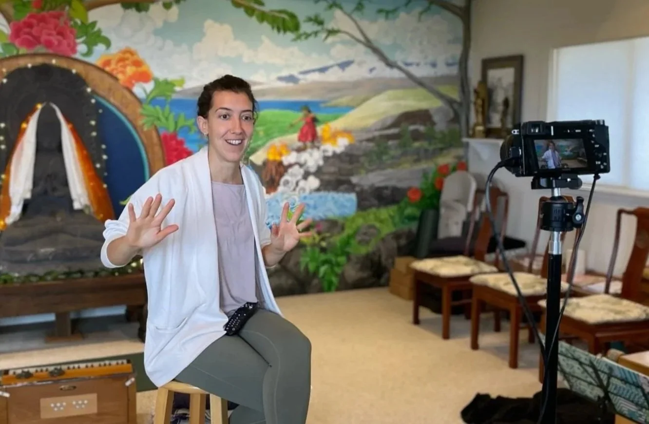 A woman sitting on a stool looks at the camera and waves, with a colorful mural depicting a landscape with a cave, a river, and a person in red in the background. A camera on a tripod records her, and there are chairs and other objects in the room.