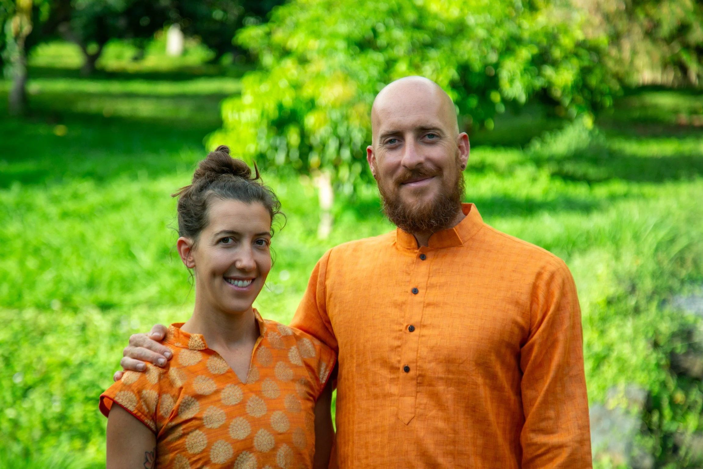 A smiling woman and man standing outdoors with green trees and grass in the background, both dressed in orange traditional clothing.