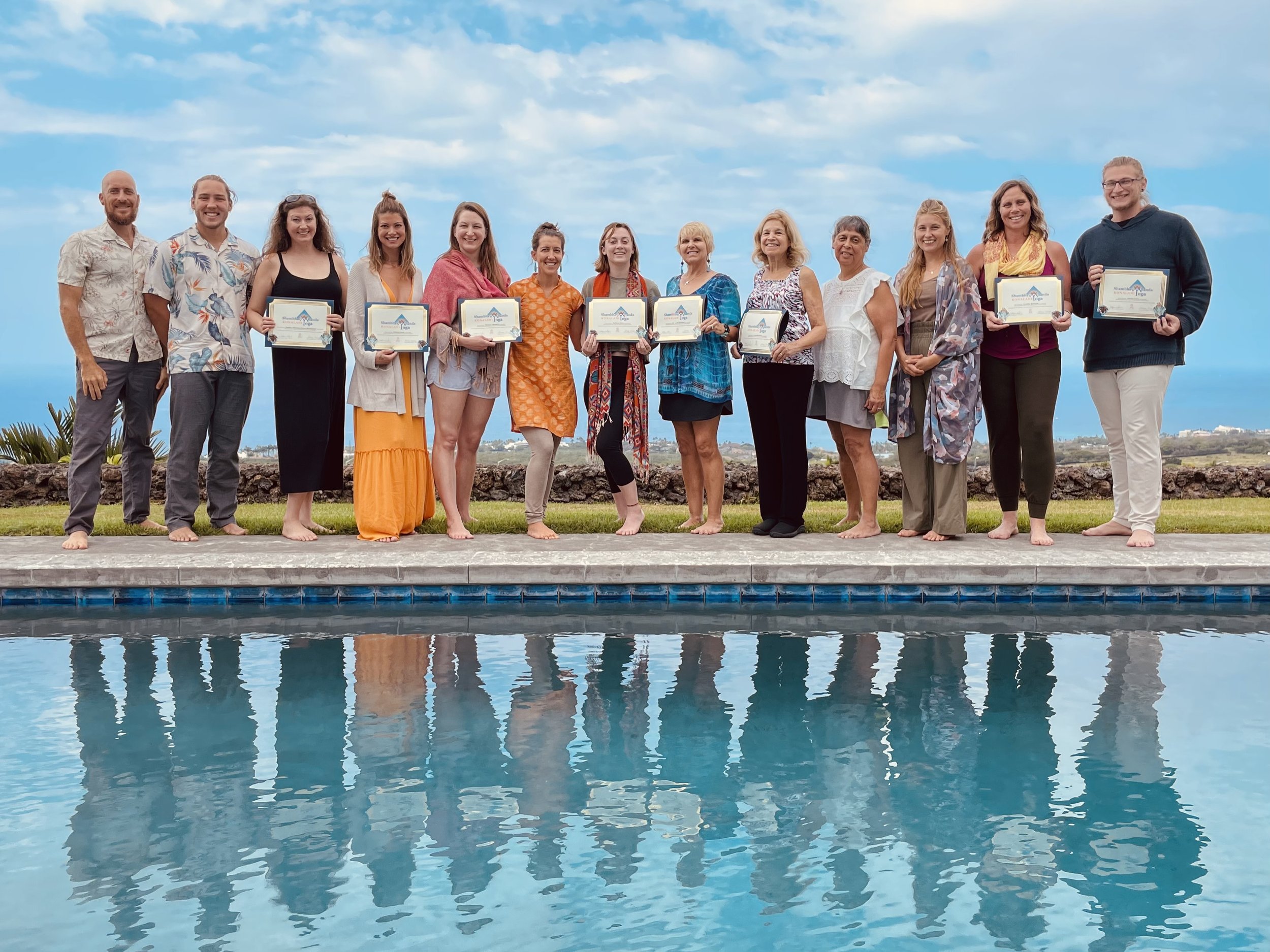 Group of 14 people standing by a pool holding certificates, outdoors with a cloudy sky in the background.