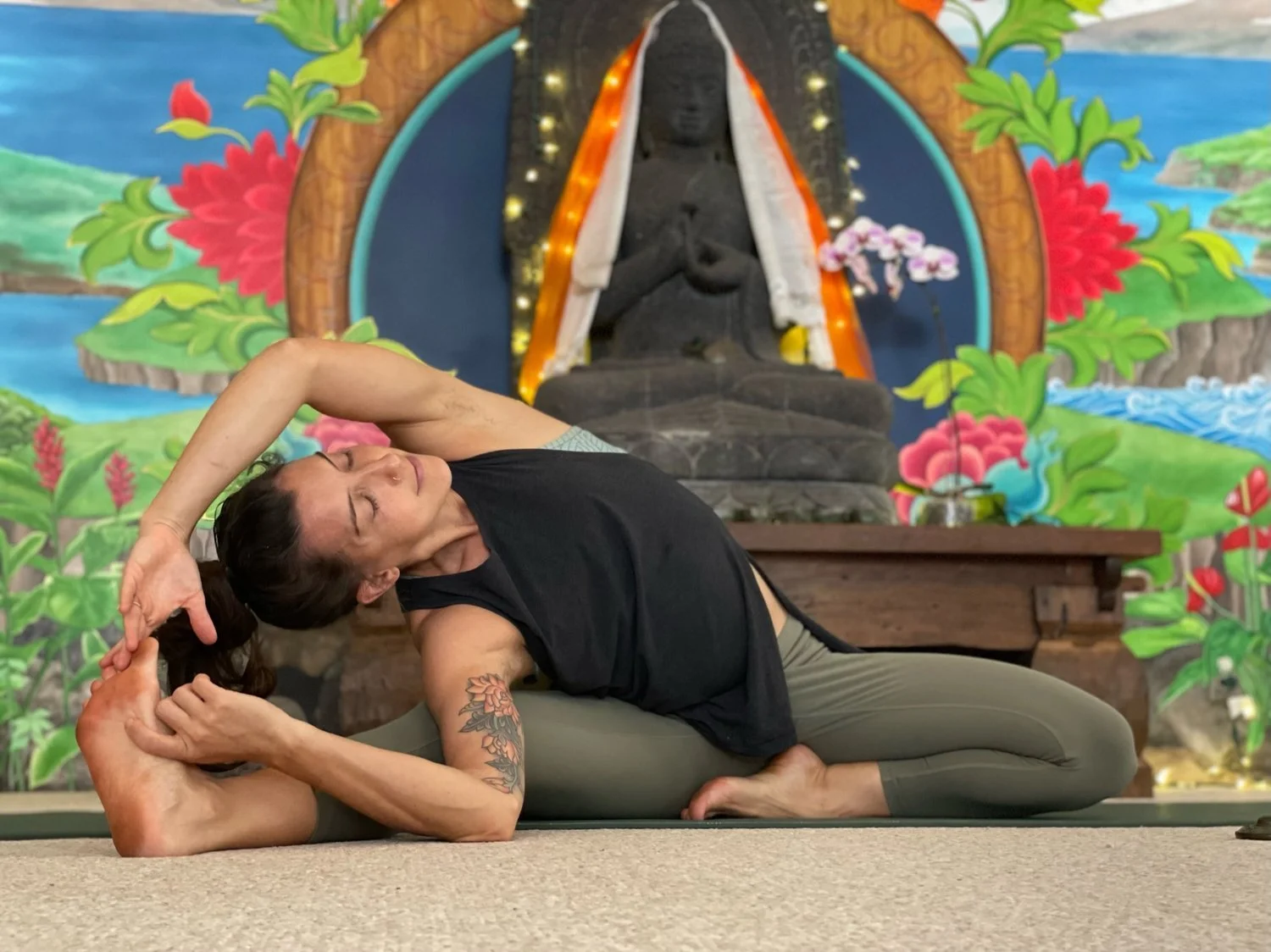 A woman practicing a yoga pose on a yoga mat in front of a colorful religious shrine featuring a seated black statue, flowers, and decorative elements.