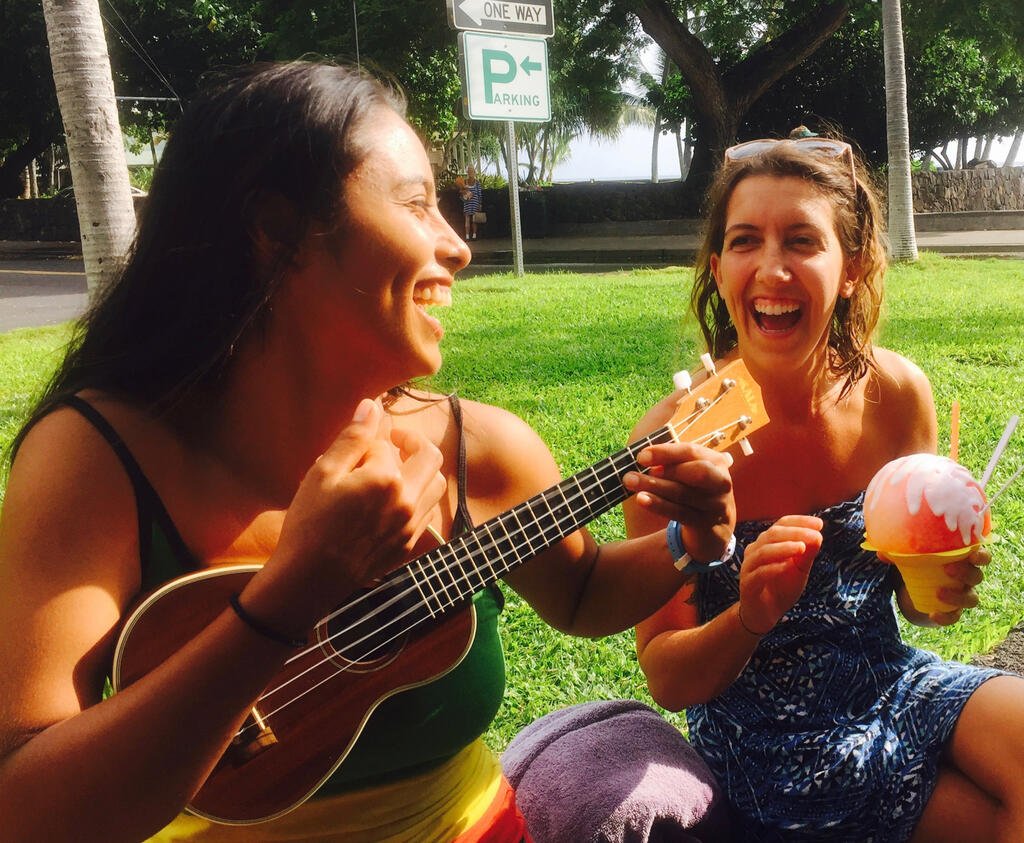 Two smiling ladies, one playing a ukulele, sit on the grass.