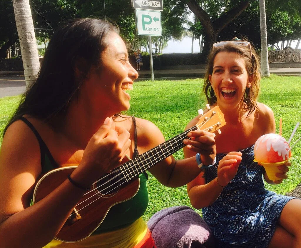 Two smiling ladies, one playing a ukulele, sit on the grass.