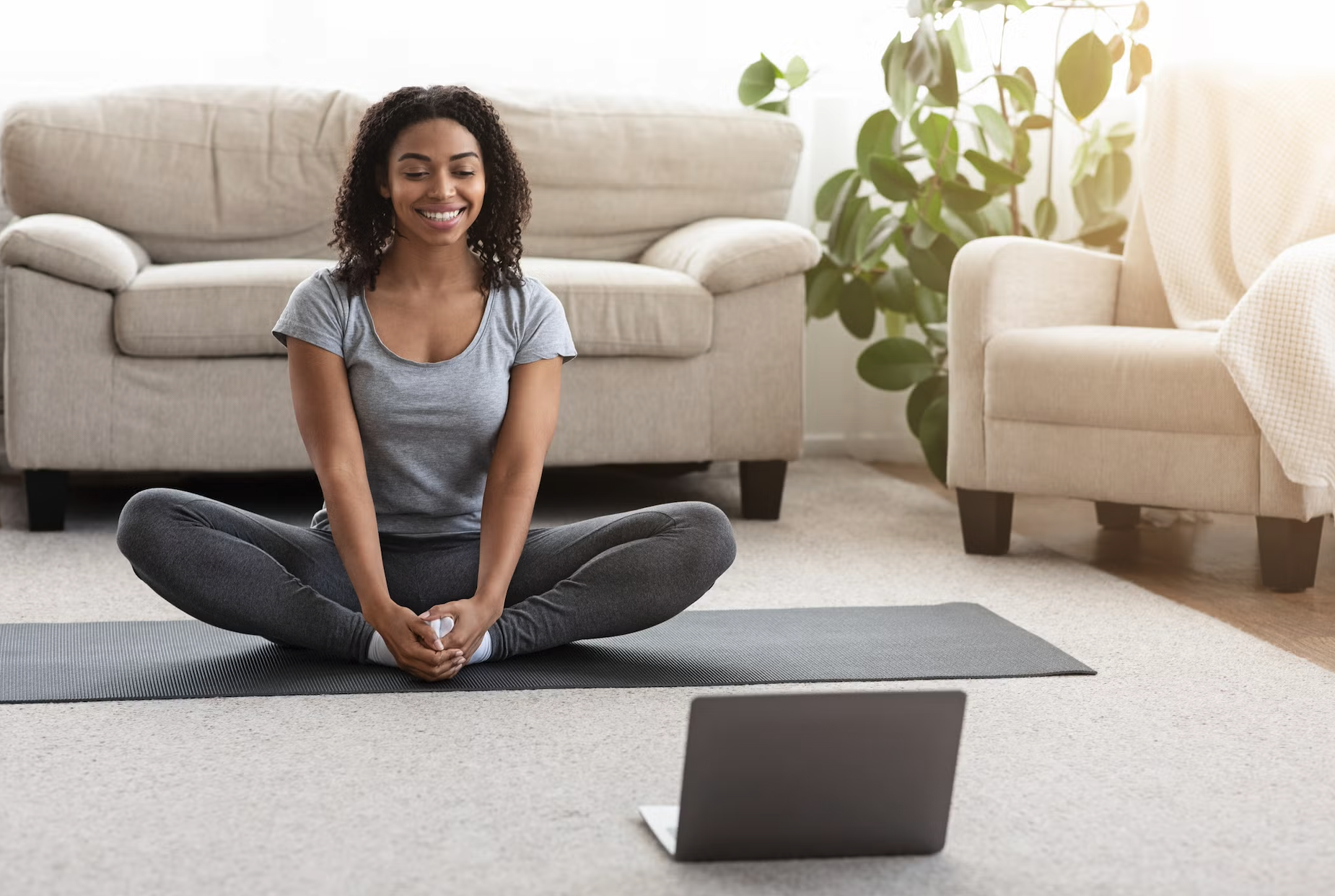 A woman sitting cross-legged on a yoga mat in her living room, smiling and engaging in a virtual yoga class on her laptop.