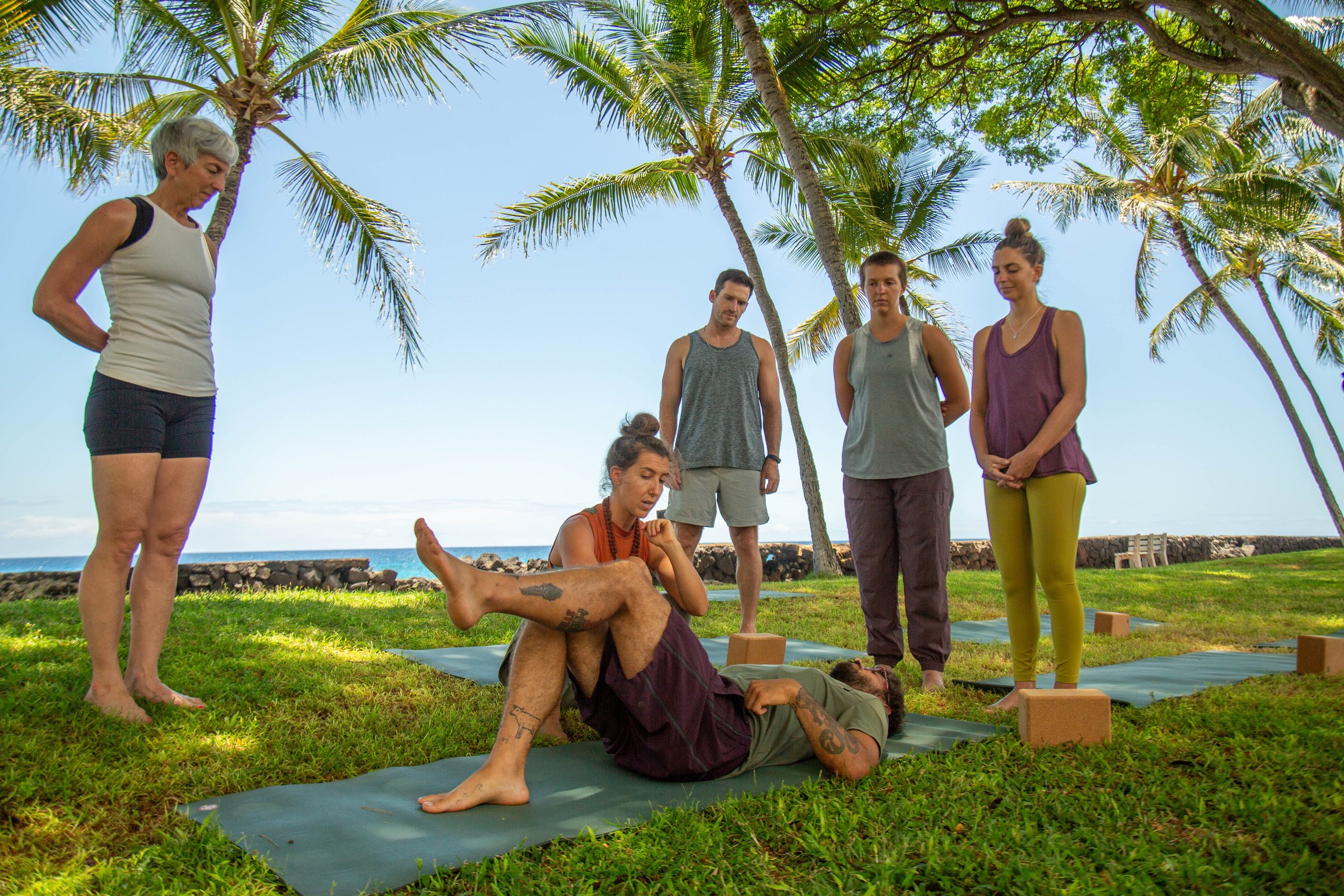 Yoga students observing a teacher demonstrate a reclining pose beside the ocean.