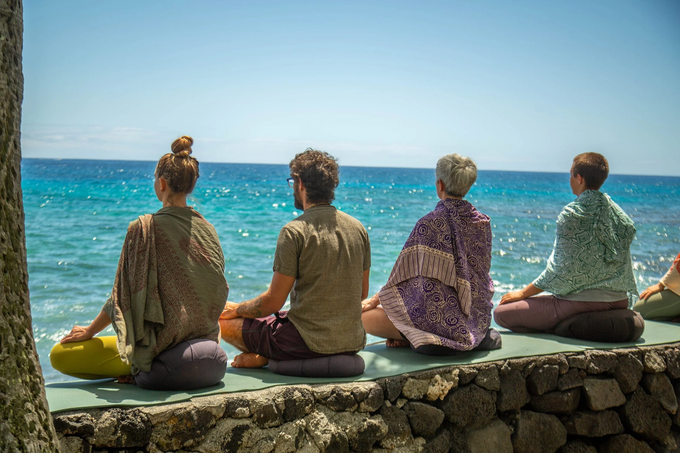 Four people sitting on a ledge by the ocean, practicing meditation on a sunny day.