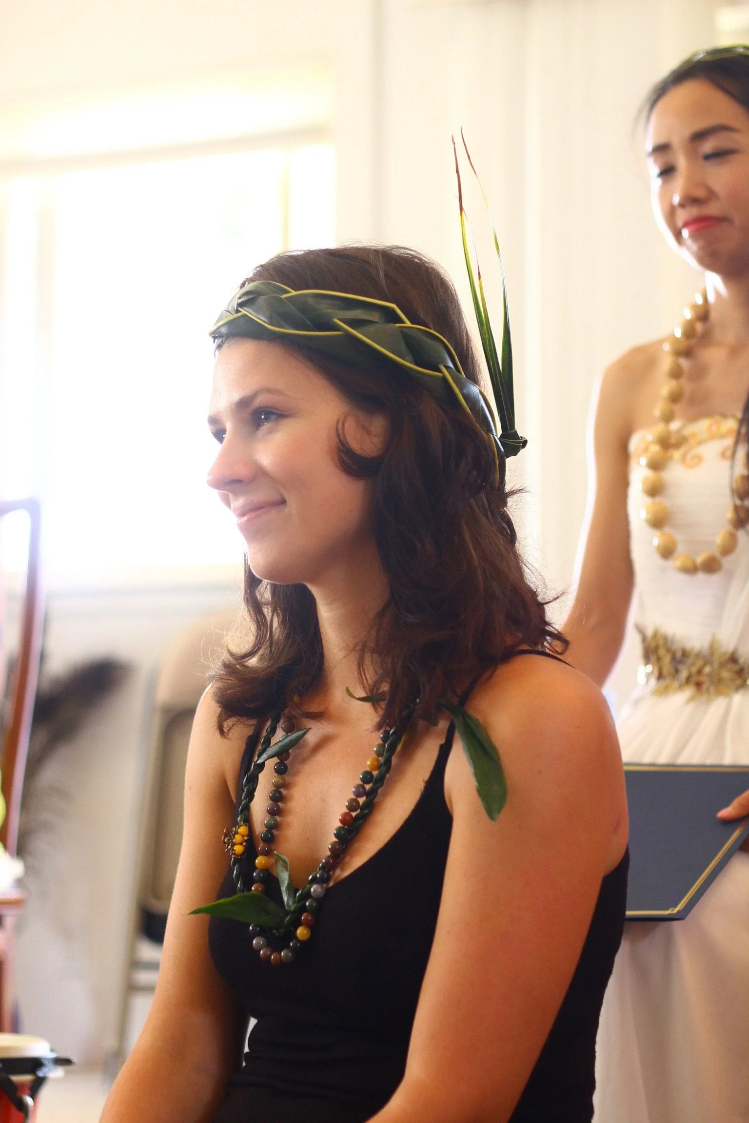 A woman with brown, curly hair wearing a black dress and a necklace, sitting indoors. Another woman in a white dress and a large necklace is standing behind her, with a large green leaf and other natural elements in her hair.