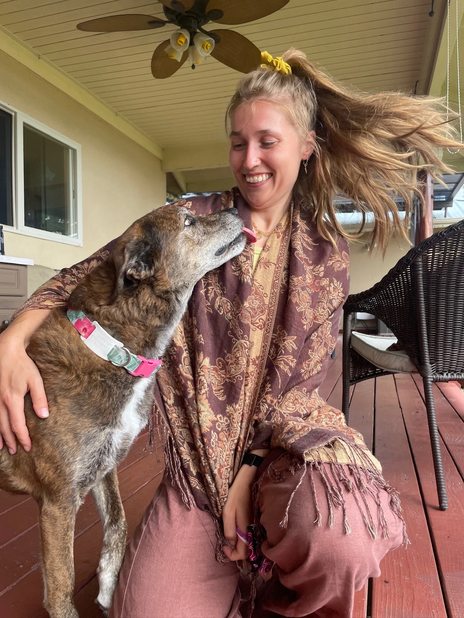 A young woman with flowing hair smiling while a dog gently licks her face, sitting on a porch with wooden floorboards, wicker chairs, and a ceiling fan overhead.