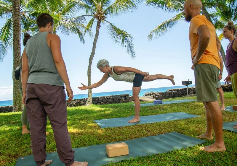 An older female yogi balances on one leg with an arm extended in front of her and a leg behind.