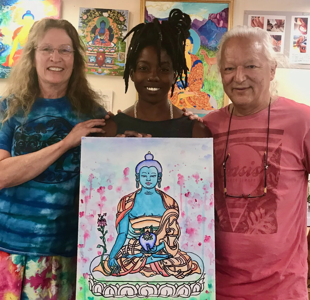 Three people standing together indoors, holding a painting of a blue-skinned Buddha figure with a pink splash background. The woman on the left has long curly hair and glasses, wearing a blue tie-dye shirt. The woman in the middle has dark hair in dreadlocks, wearing a black top and a pearl necklace. The man on the right has white hair, wearing a pink shirt and glasses hanging around his neck. Artworks are displayed on the wall behind them.