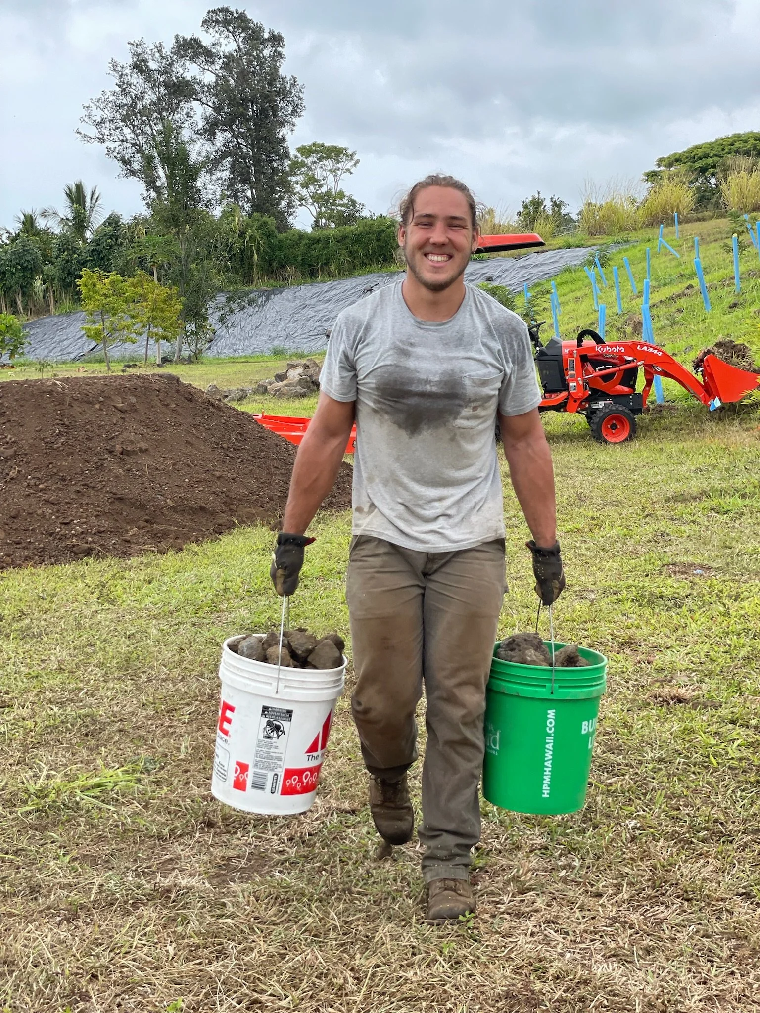 A smiling man wearing work gloves and muddy clothes, carrying two buckets filled with rocks, in a grassy outdoor area with trees and construction equipment in the background.