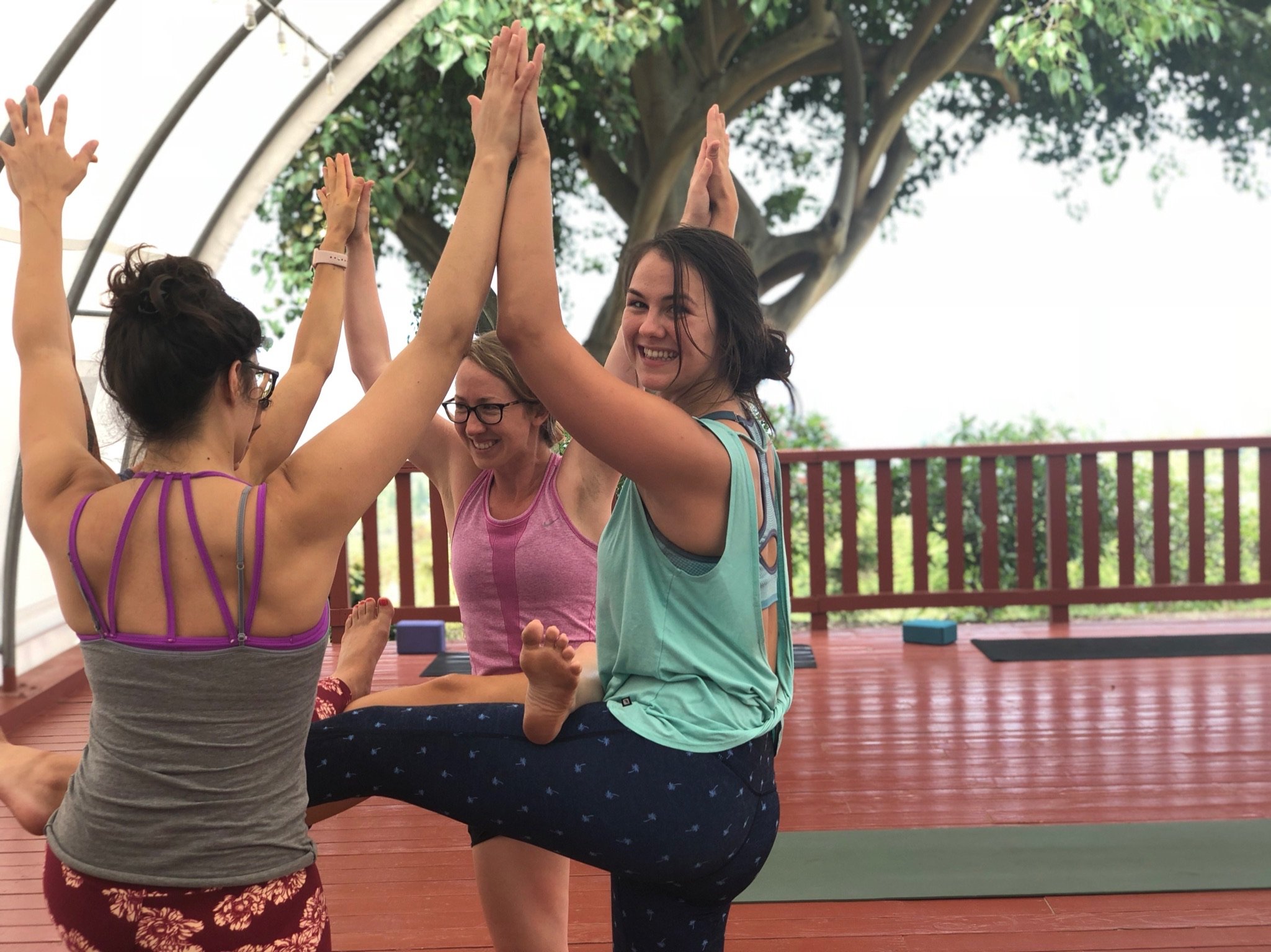 Three woman yogis practice partner yoga touching hands and legs while smiling toward the camera on a covered deck.