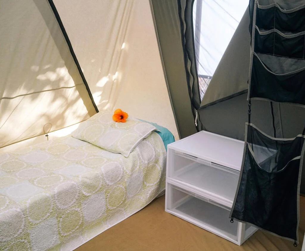 A nicely made single cot with an orange flower on the pillow next to plastic storage container in a large tent.