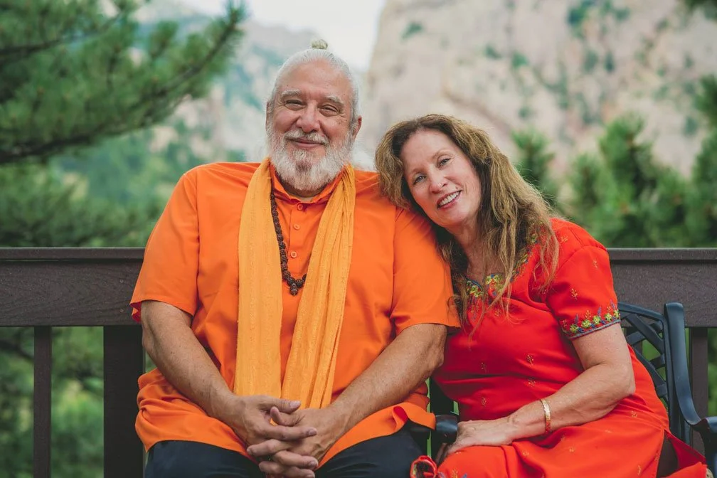 A smiling elderly man with a white beard and a woman, both wearing orange outfits, sitting on a bench outdoors with green trees in the background.