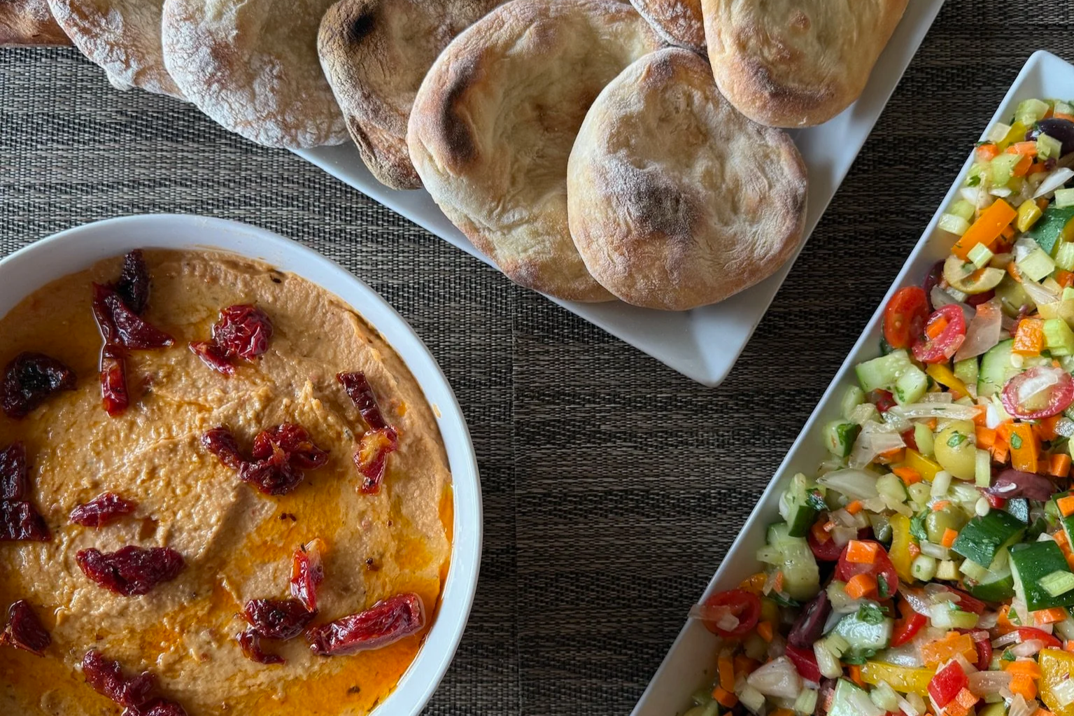 Plate of hummus with sun-dried tomatoes, assorted bread rolls, and a colorful chopped vegetable salad on a dark textured table.