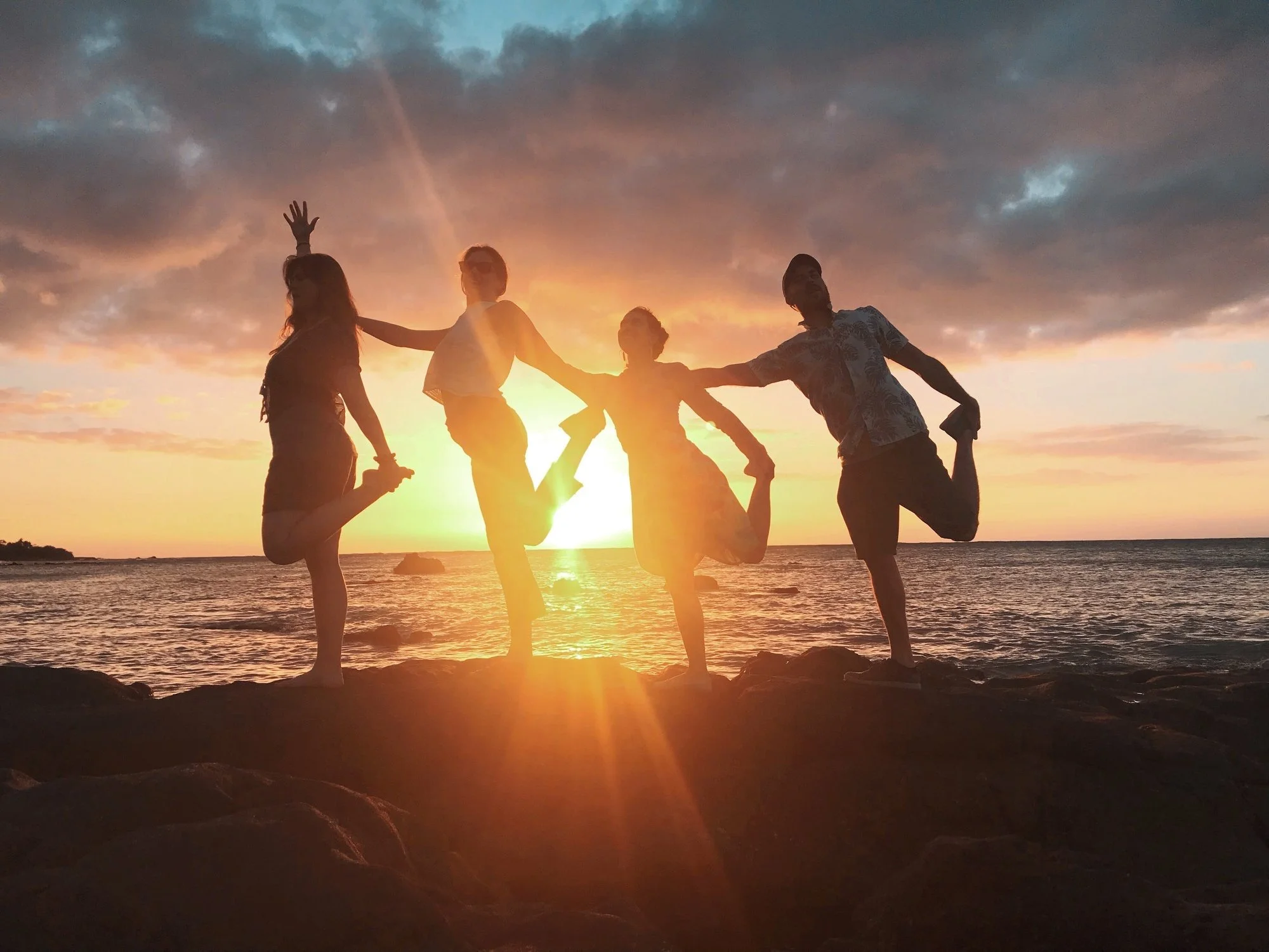 Four friends balancing together beside the ocean at sunset.