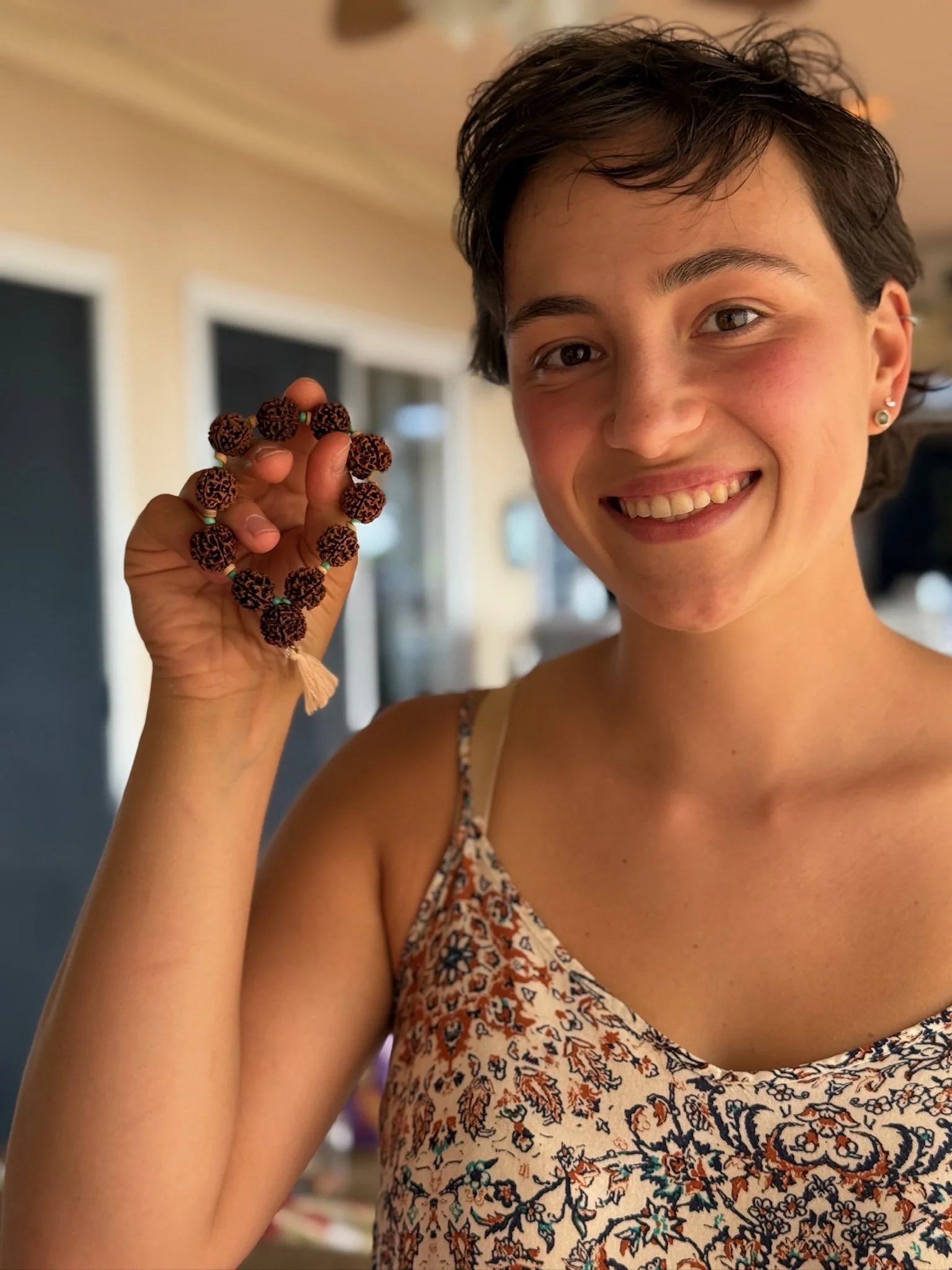 Smiling young woman with short dark hair wearing a sleeveless floral top, holding a loyalty bracelet made of large dark beads with small beads in between, indoors with windows and blurred background.
