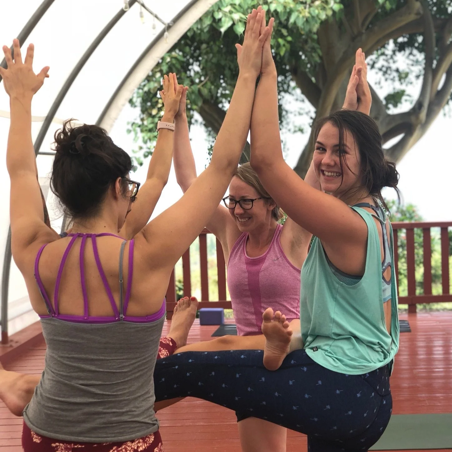 Four women doing acro yoga outside on a deck, smiling and high-fiving each other, with a large tree and cloudy sky in the background.