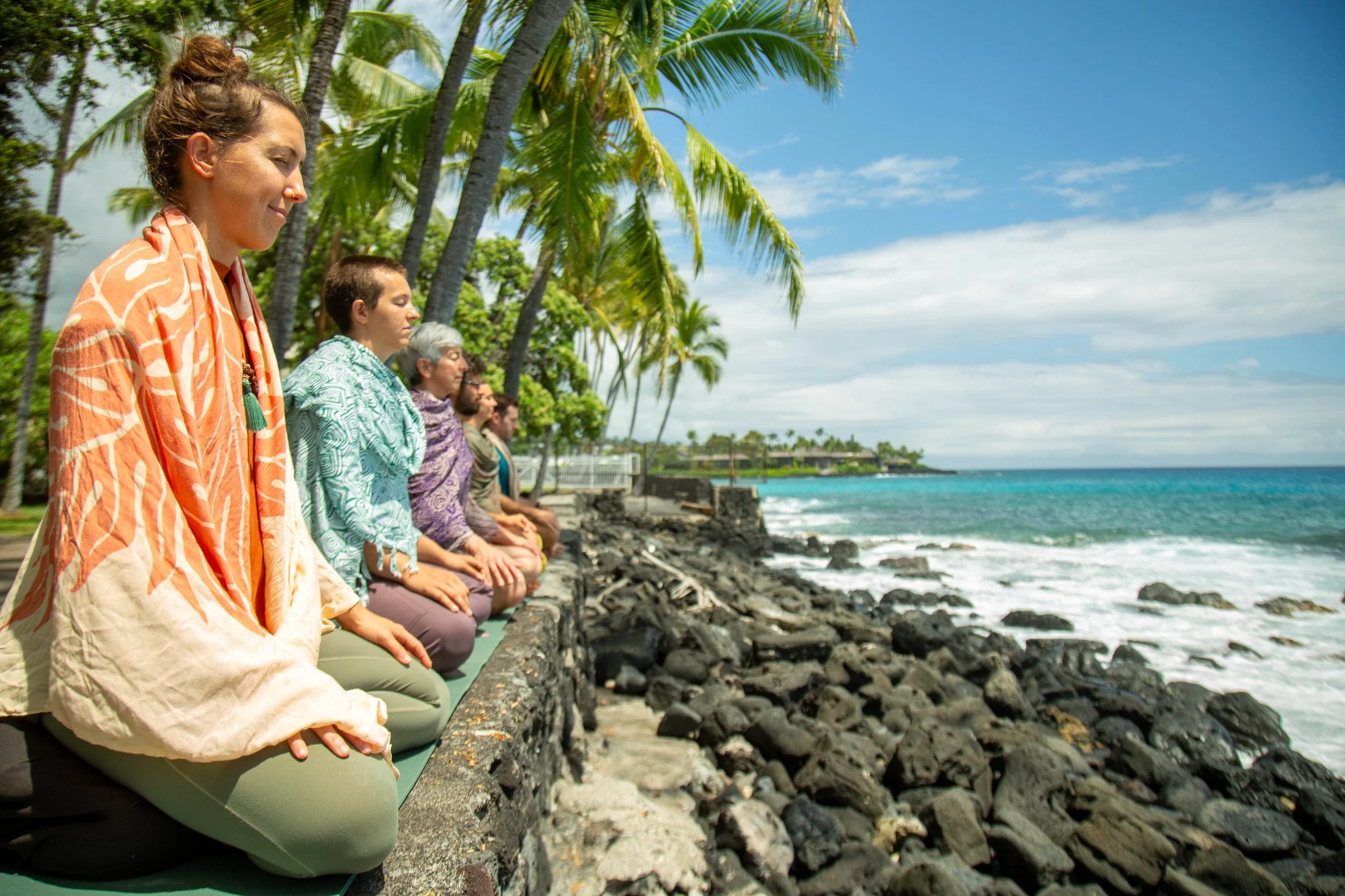 A group of people meditating outdoors on a pathway by the ocean, sitting with their legs crossed and hands resting on their laps, surrounded by palm trees with a view of the rocky shoreline and blue sky.
