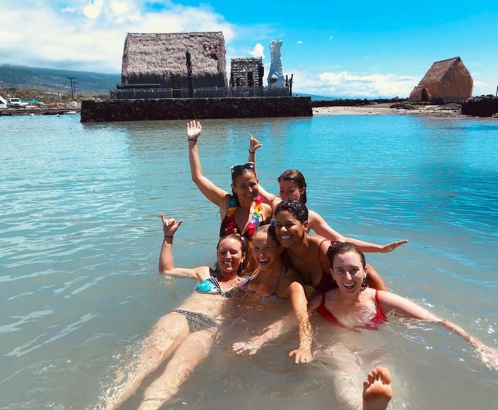 A group of swimmers raise their hands to the sky in a protected bay with historical structures in the background.