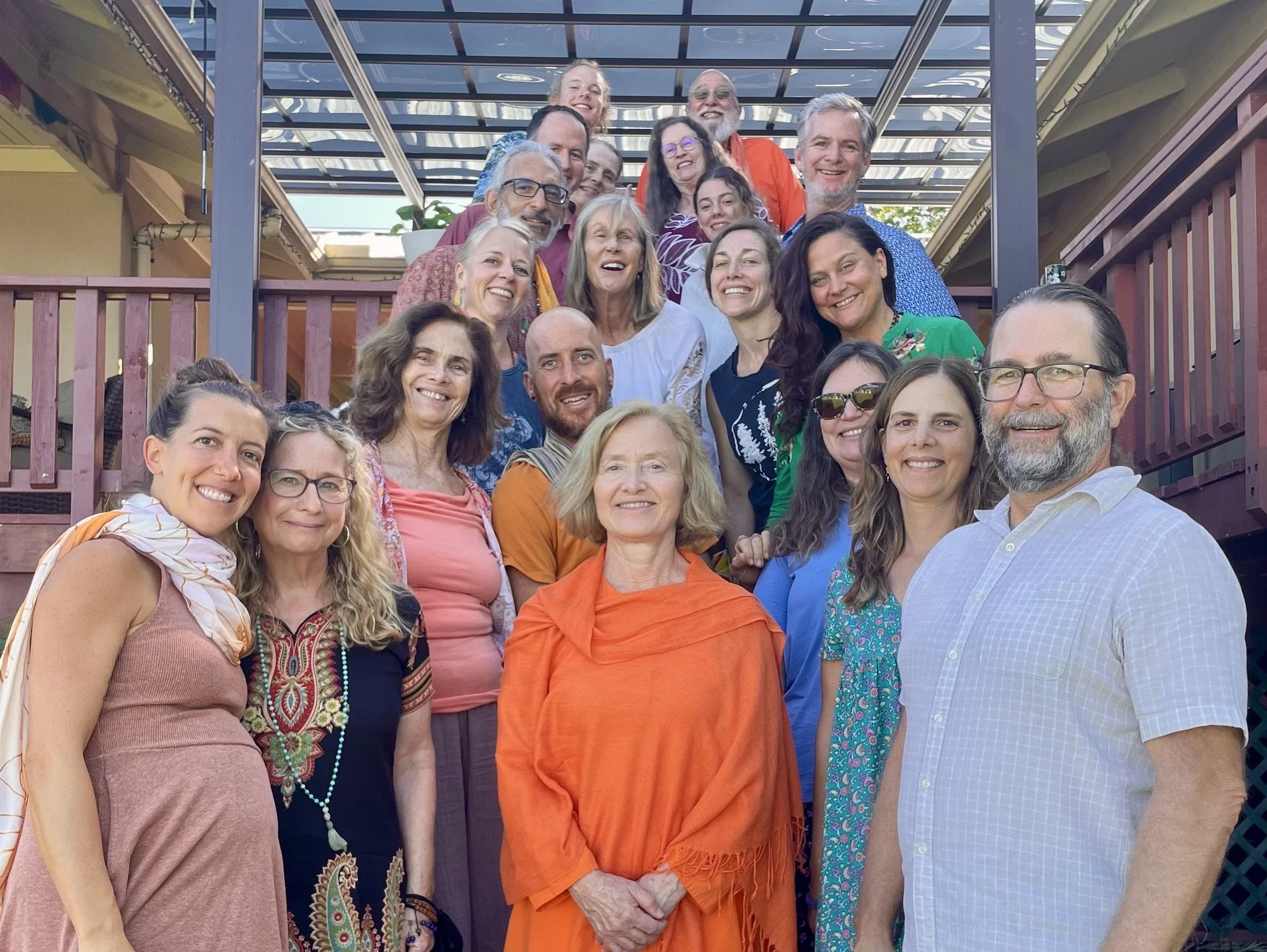 A group photo of 17 people standing on and in front of a deck, smiling and posing outdoors under a glass roof with wooden railings, in sunny weather.